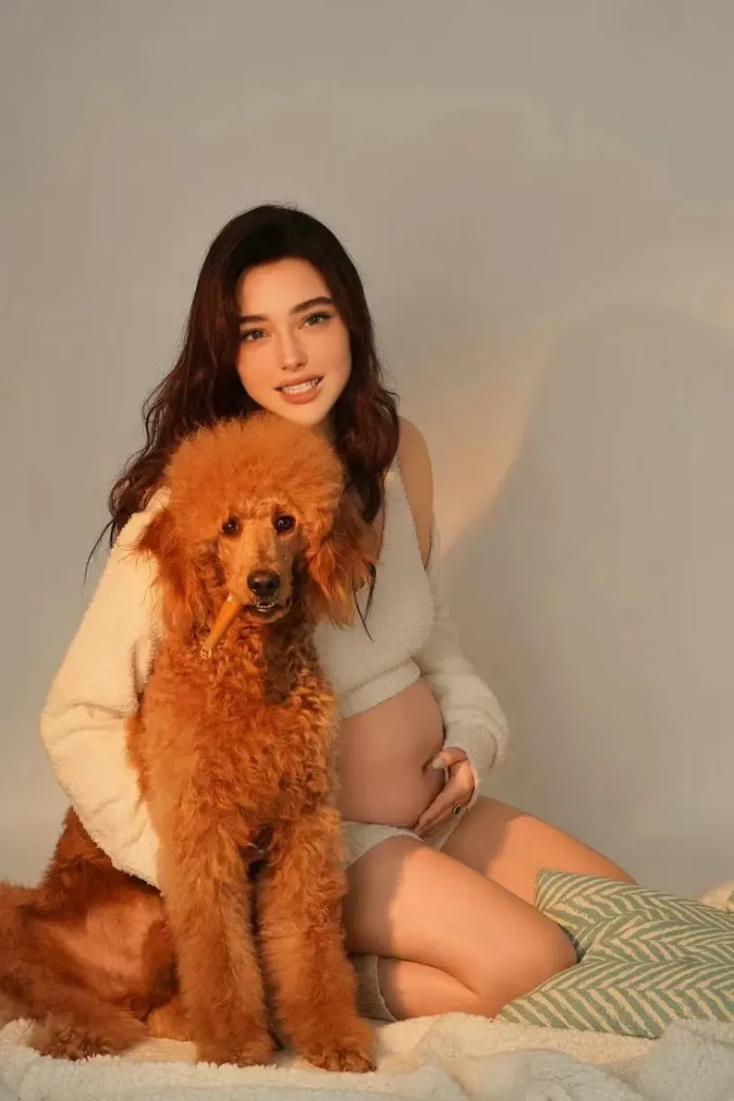 Smiling pregnant woman in cream knit outfit sitting on bed with large golden red poodle dog.