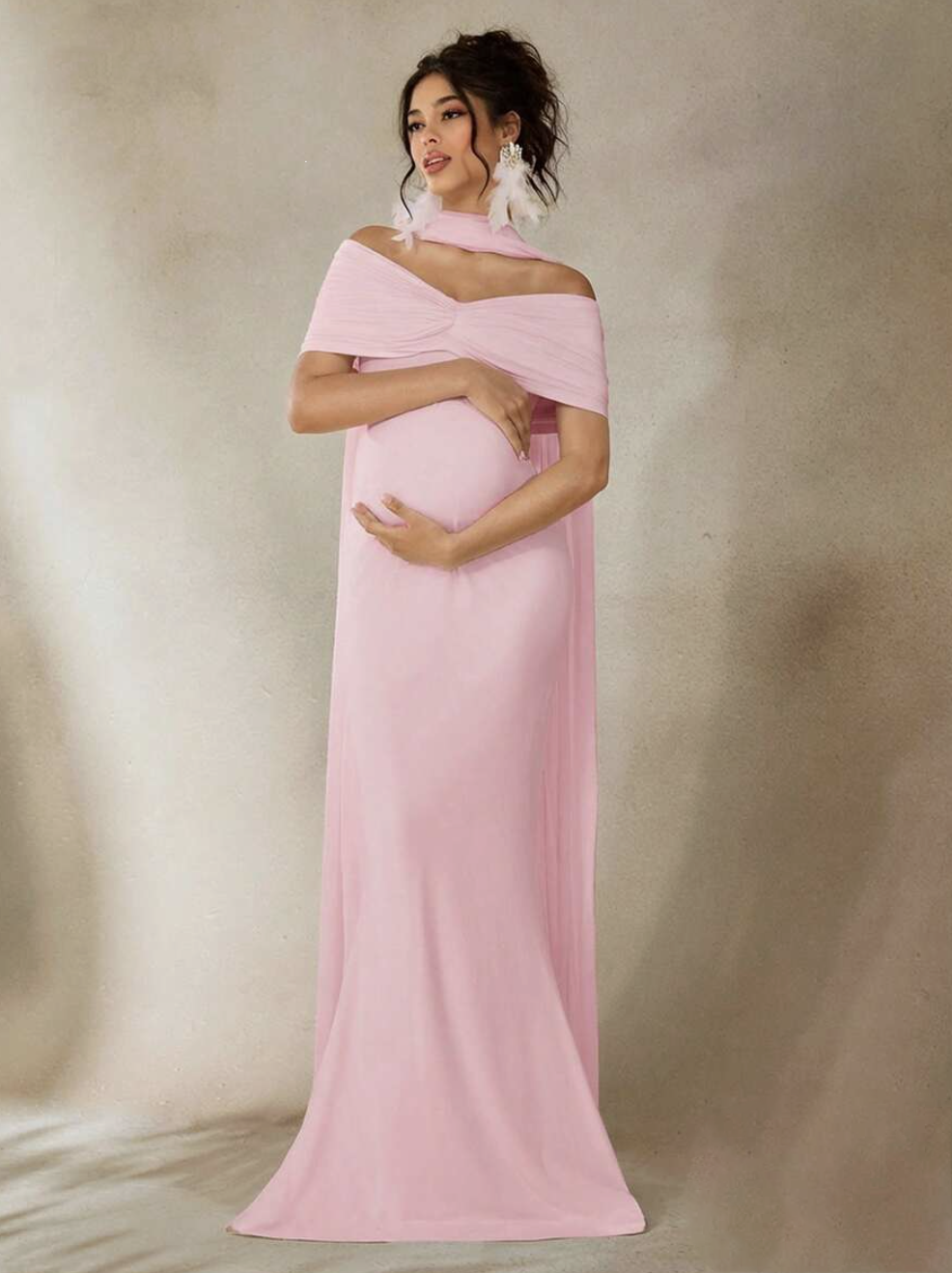 Pregnant woman wearing a pink off-shoulder maxi dress, posing elegantly with white feather earrings.