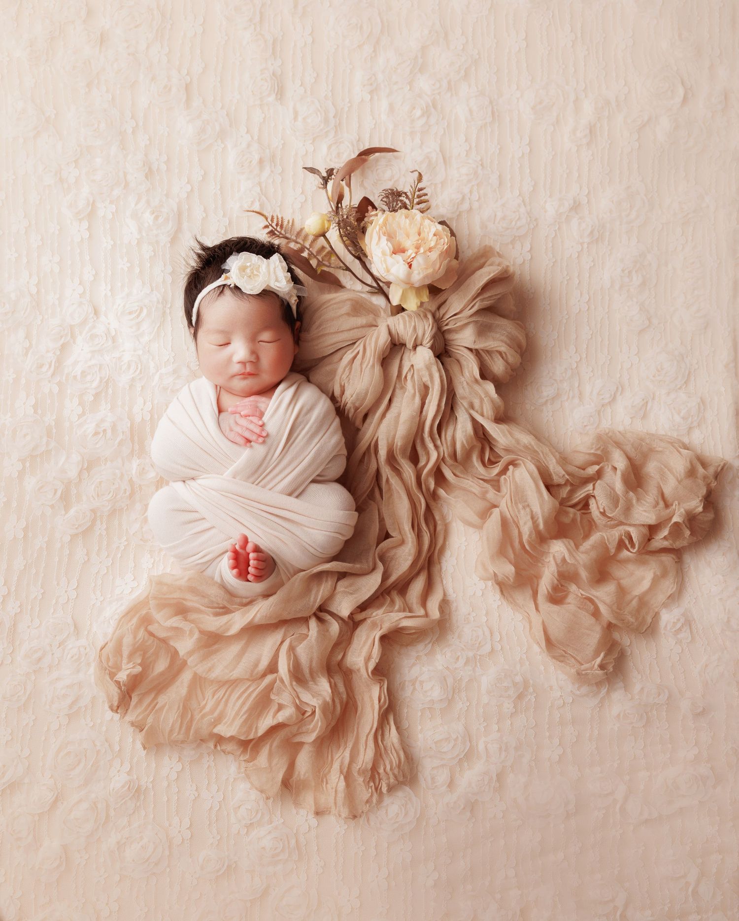 Swaddled newborn baby girl with white flower headband on beige textured wrap.