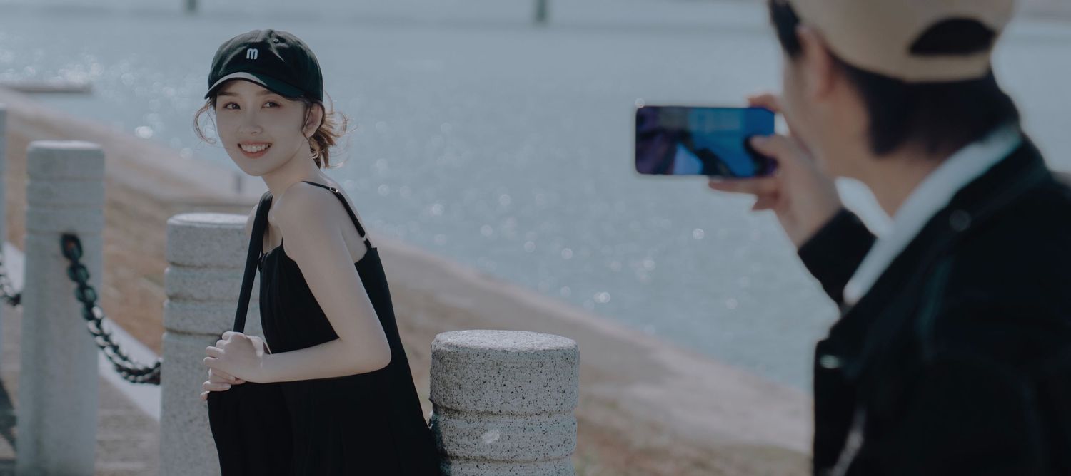 A young woman in a black dress smiling while a man photographs her near a sparkling waterfront.