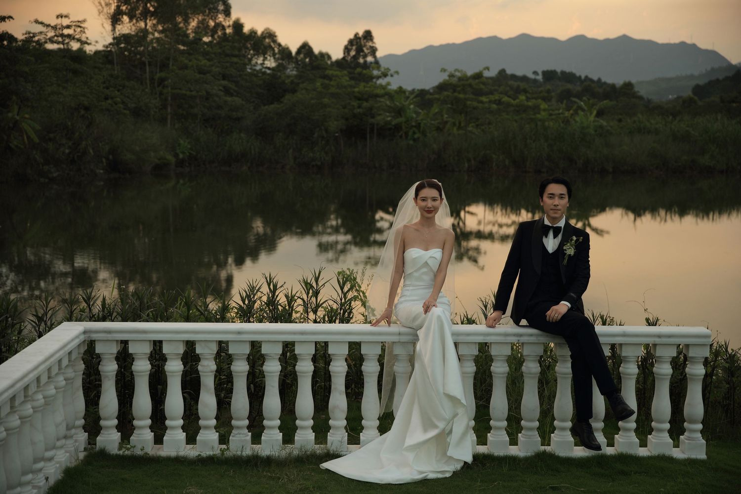 Wedding couple poses on white balustrade terrace overlooking tranquil lake at golden sunset.