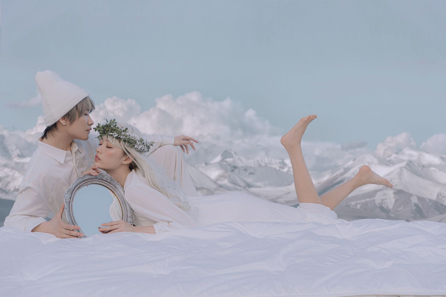 Dreamy couple in white outfits lying on snowy ground with a mirror in a winter fantasy setting.