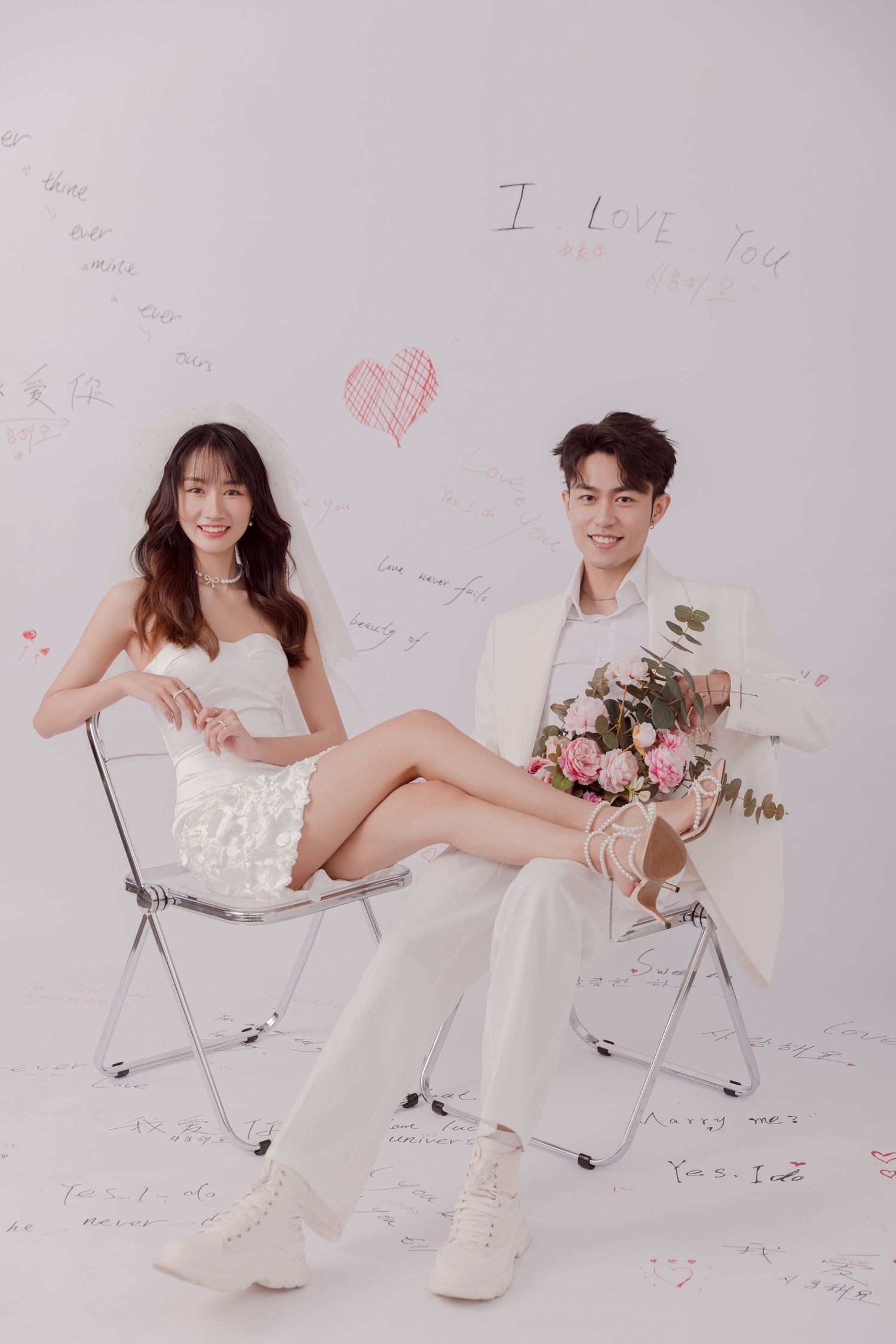 Stylish couple in white outfits sitting on chairs before a wall covered in romantic love messages.