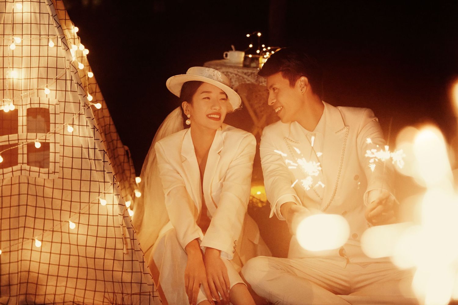 Wedding couple in white outfits sit together holding sparklers surrounded by warm string lights at night.