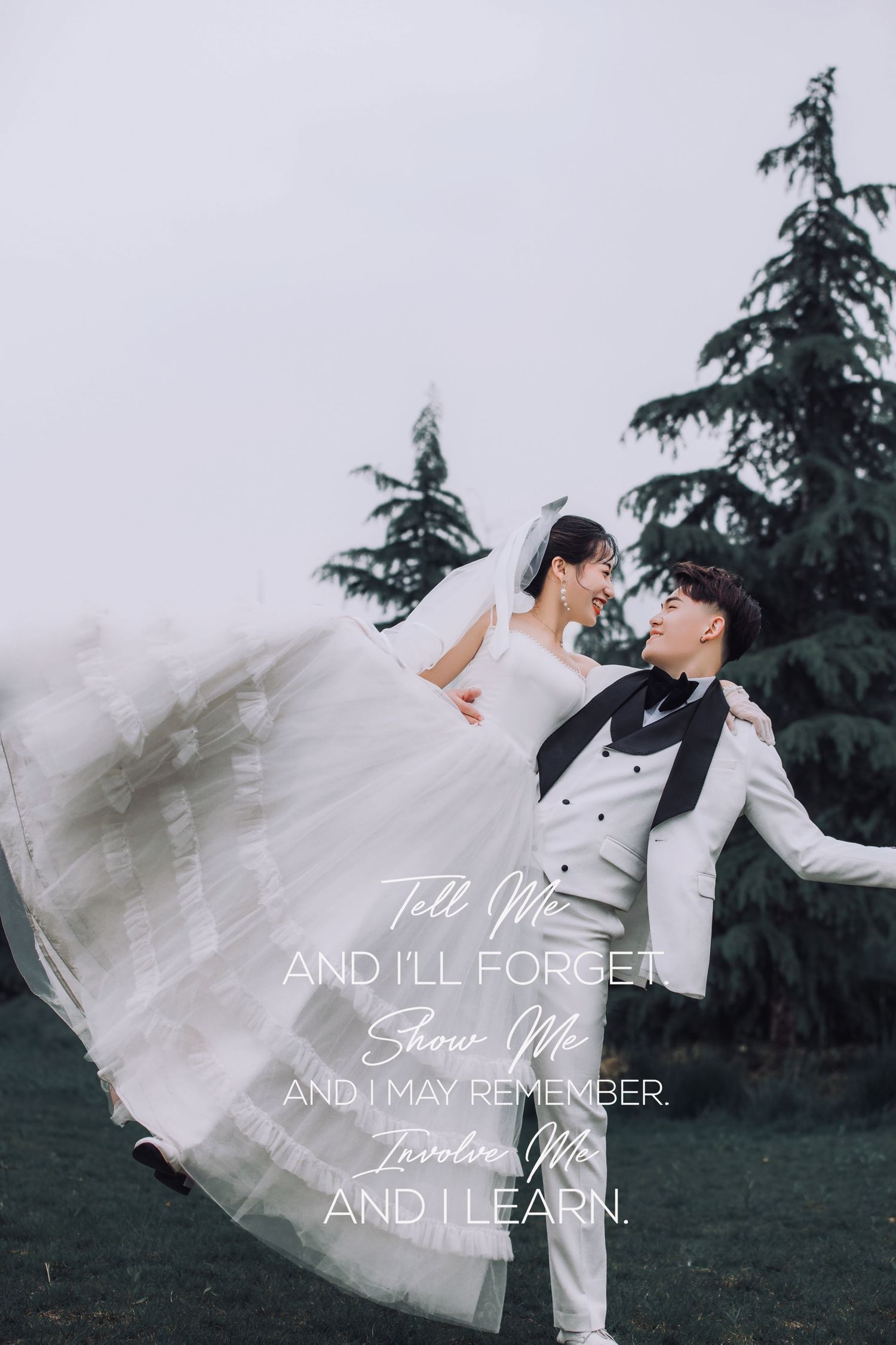 Joyful groom lifts bride in flowing white wedding dress outdoors with evergreen trees backdrop.
