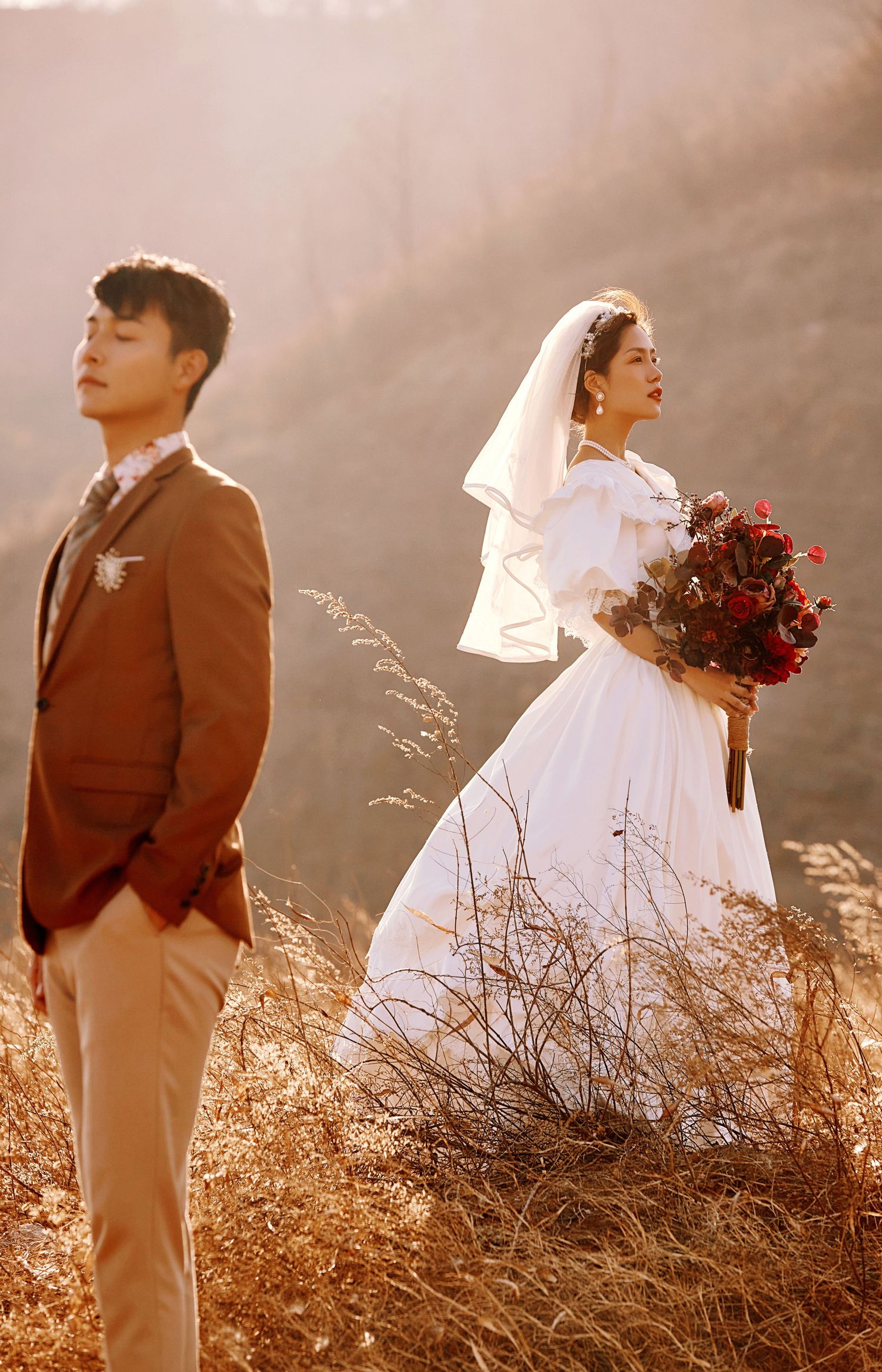 Bride in white puff-sleeve gown holds red bouquet beside groom in brown suit in golden meadow.