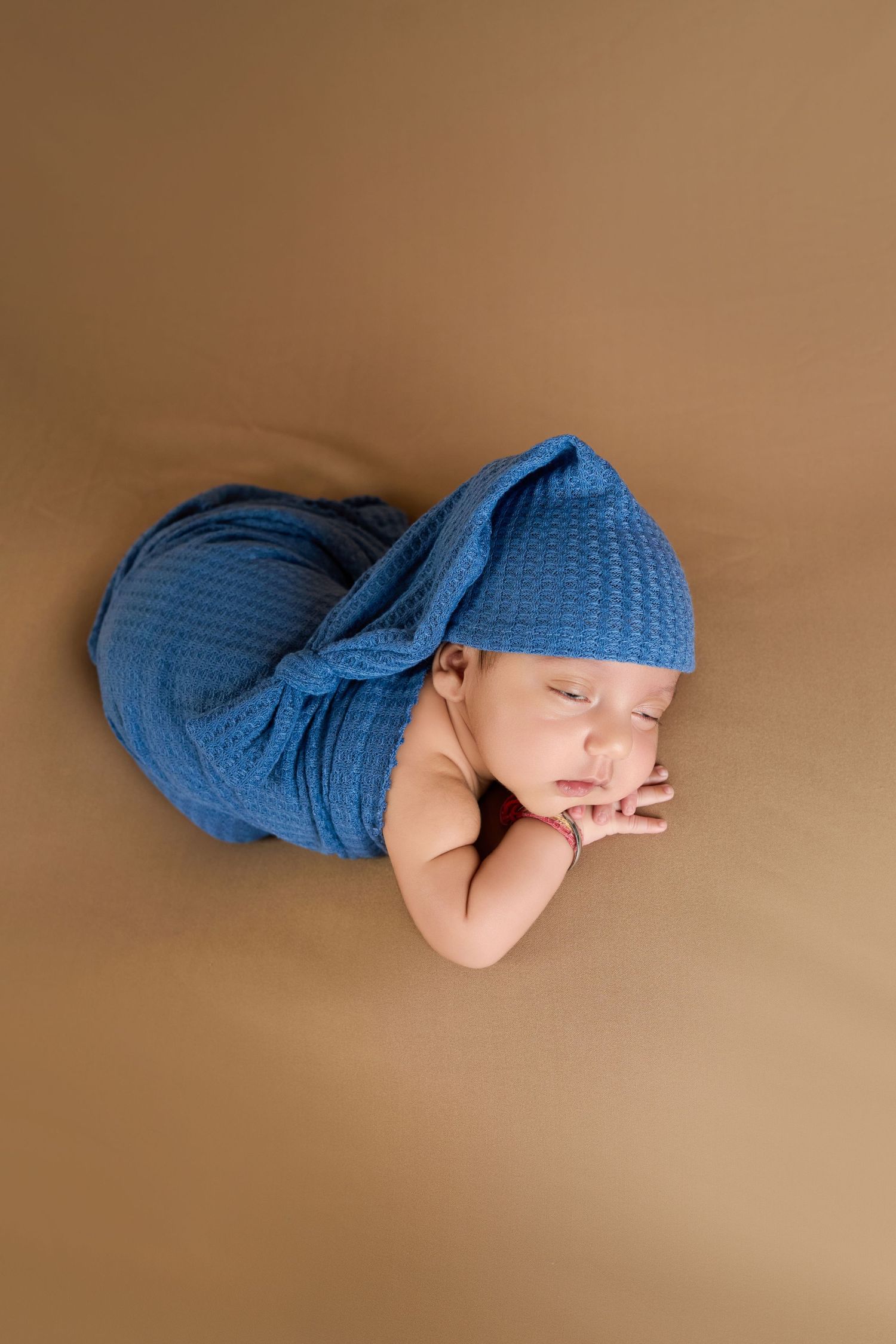 Sleeping newborn baby wrapped in a blue knit blanket and matching hat, posed on a tan background.