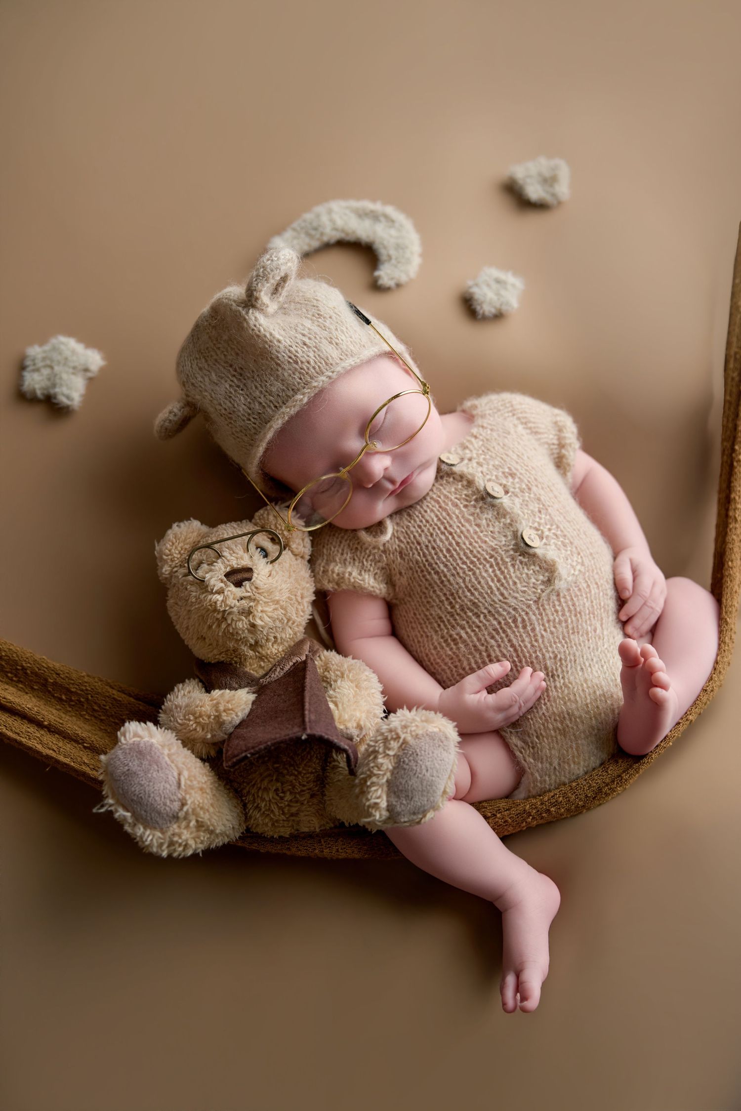 Newborn baby sleeping in a hammock wearing a knit bear outfit and glasses, cuddling a teddy bear on a beige background.