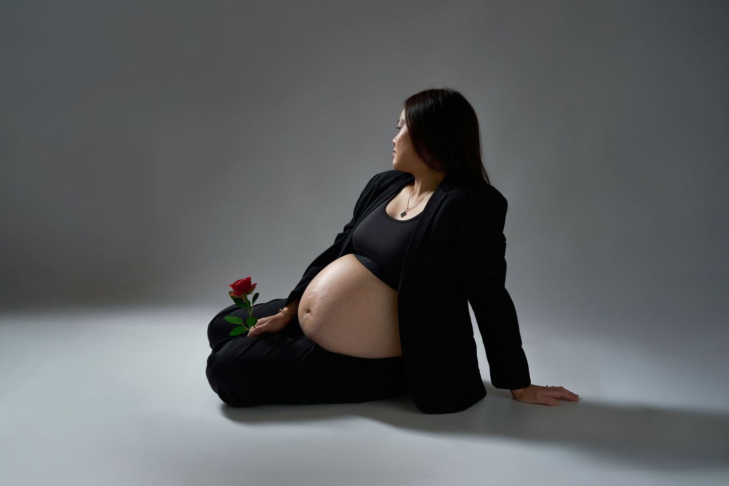 Pregnant woman in black outfit sitting on floor holding a red rose, showcasing baby bump in a studio portrait.
