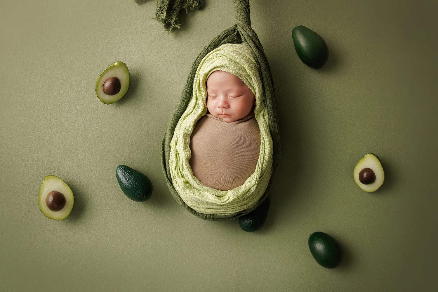 Newborn baby sleeping in avocado-shaped prop surrounded by avocados on green background.