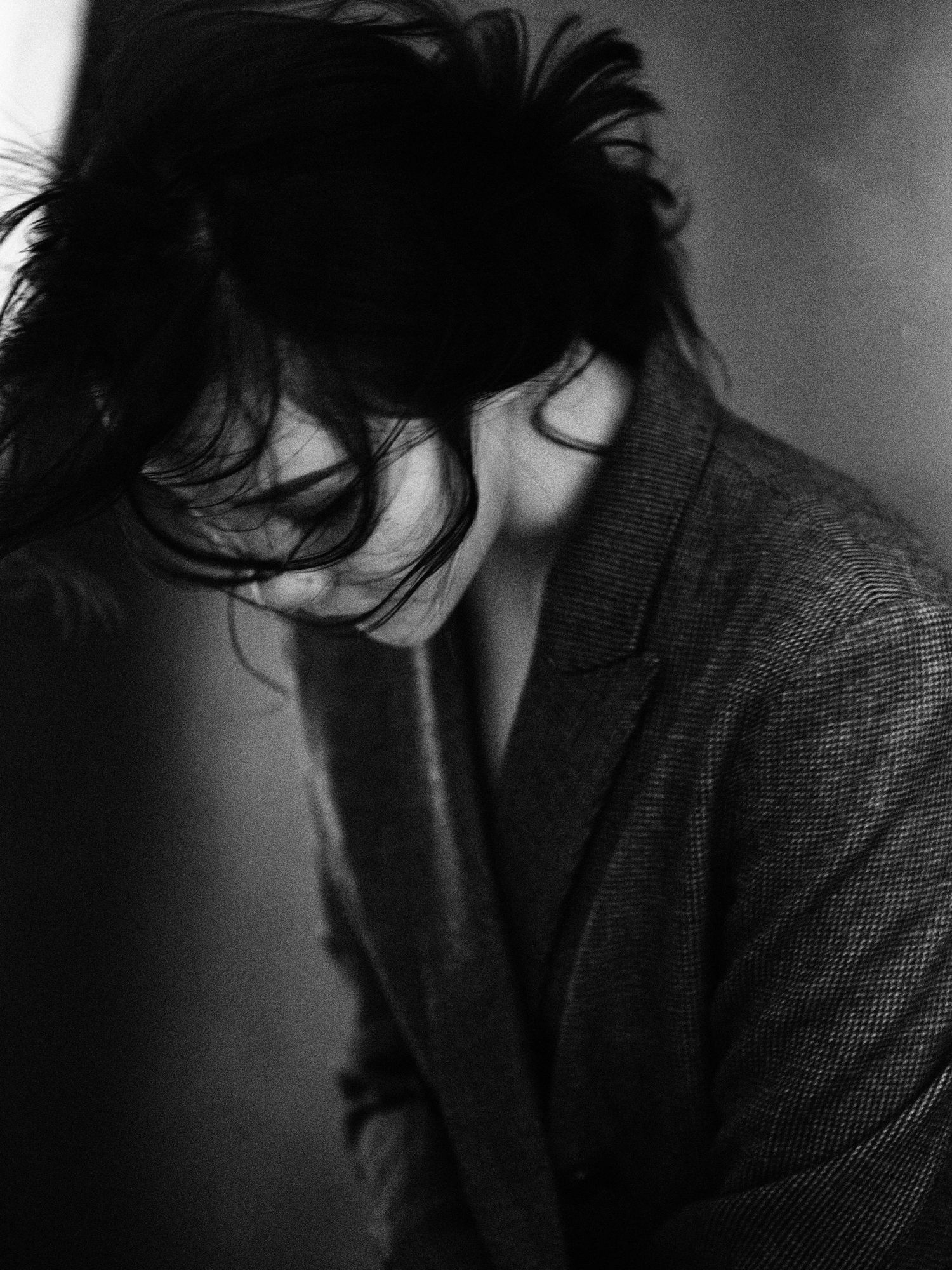 Moody black and white photo of a woman with windswept dark hair wearing an oversized blazer, head bowed down.
