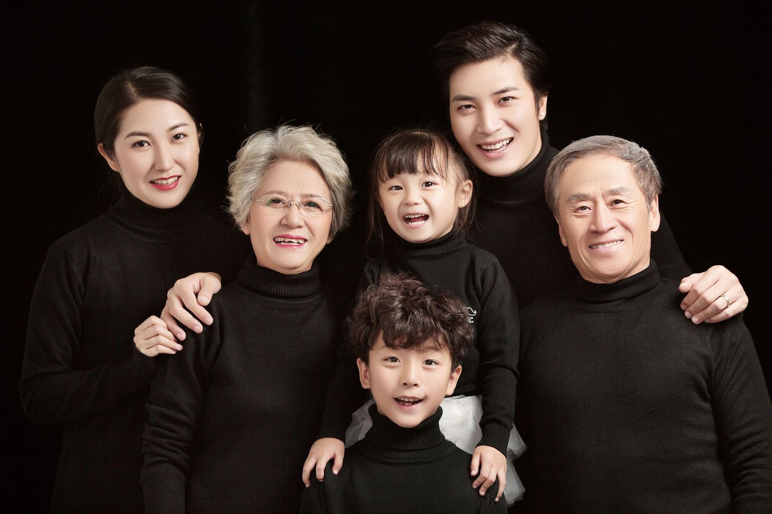 A multigenerational Asian family of six smiles together wearing matching black turtlenecks in a studio.