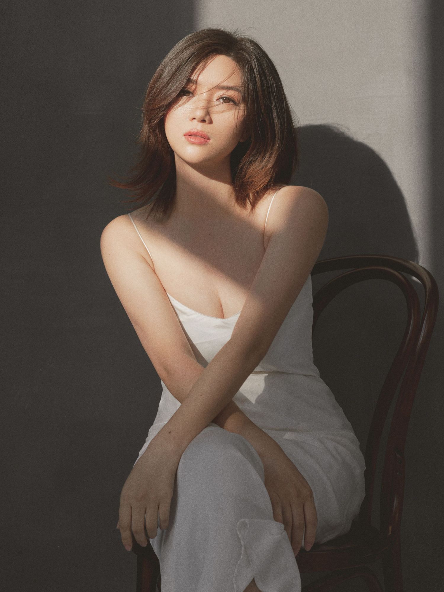 Young woman in white spaghetti-strap dress sits on wooden chair with arms crossed, lit by dramatic sunlight.