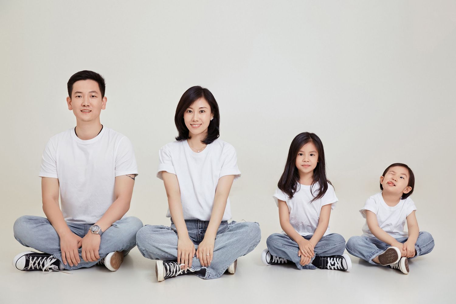 Asian family of four in matching white shirts and jeans sit cross-legged in a row in a studio.