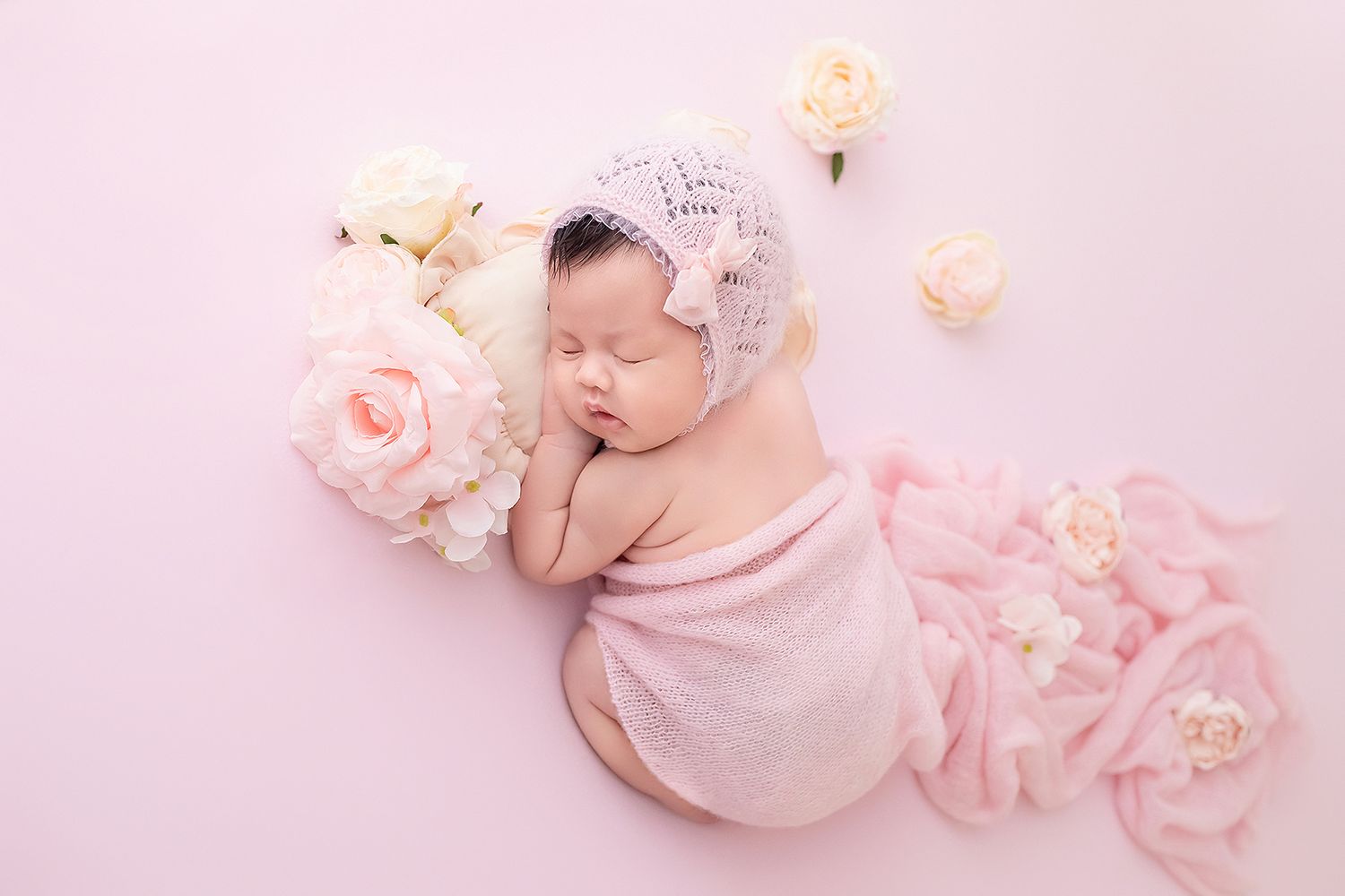 Newborn baby sleeping wrapped in pink blanket with knit bonnet hat surrounded by white and pink flowers.