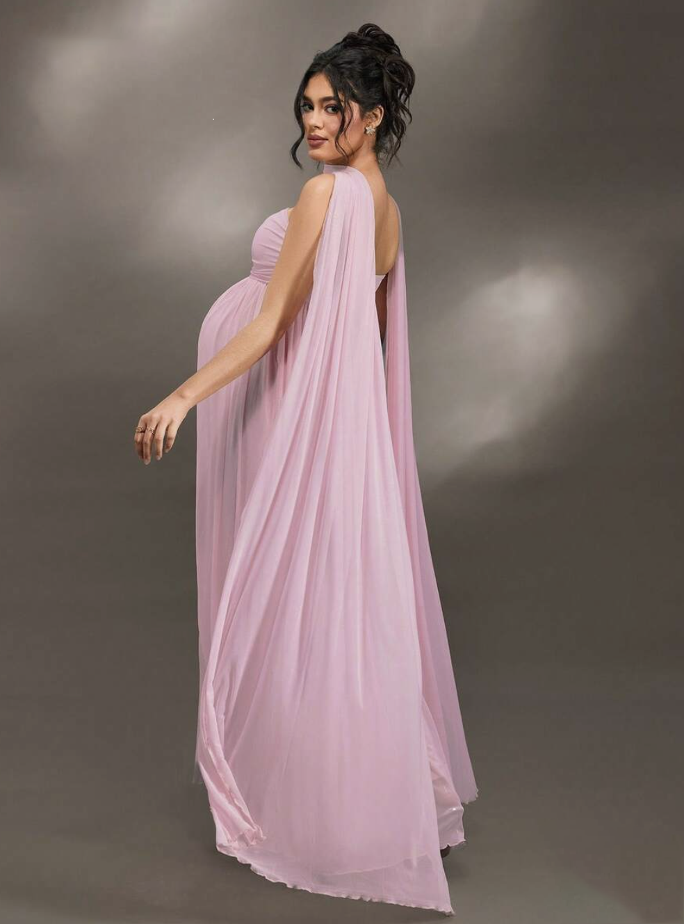 Pregnant woman in elegant pink chiffon cape maxi dress, flowing one-shoulder gown for maternity formal wear.