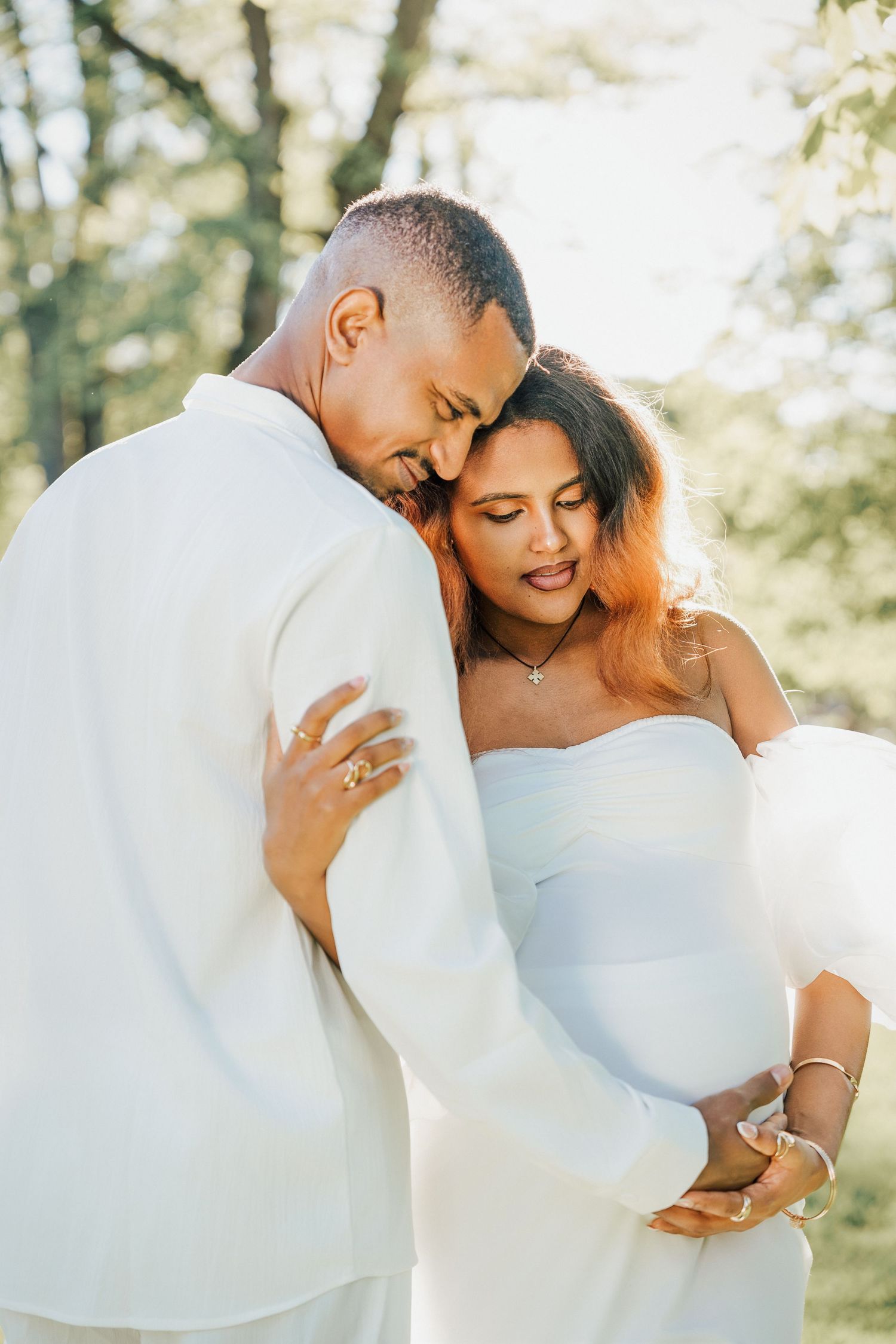 Couple in white outfits share tender moment outdoors, pregnant woman cradling baby bump while partner embraces her lovingly.