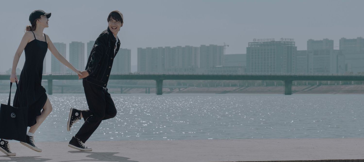 A couple runs along a waterfront promenade with city skyline in the background, pulling luggage.