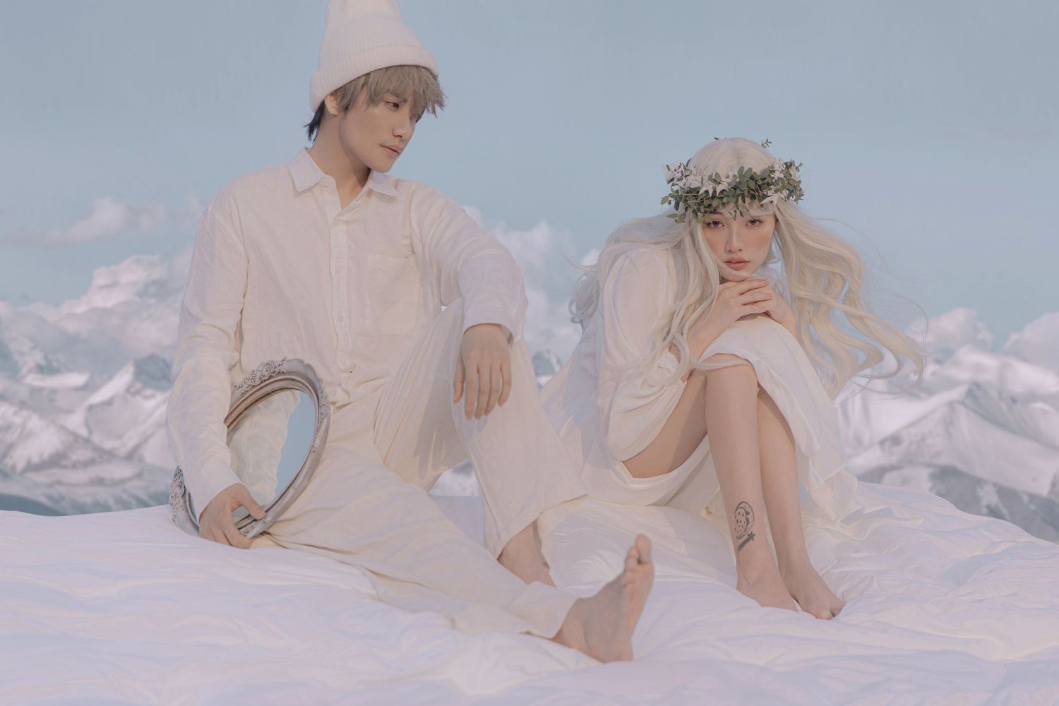 Couple in all-white outfits sit together outdoors, woman wearing flower crown, man holding silver mirror.
