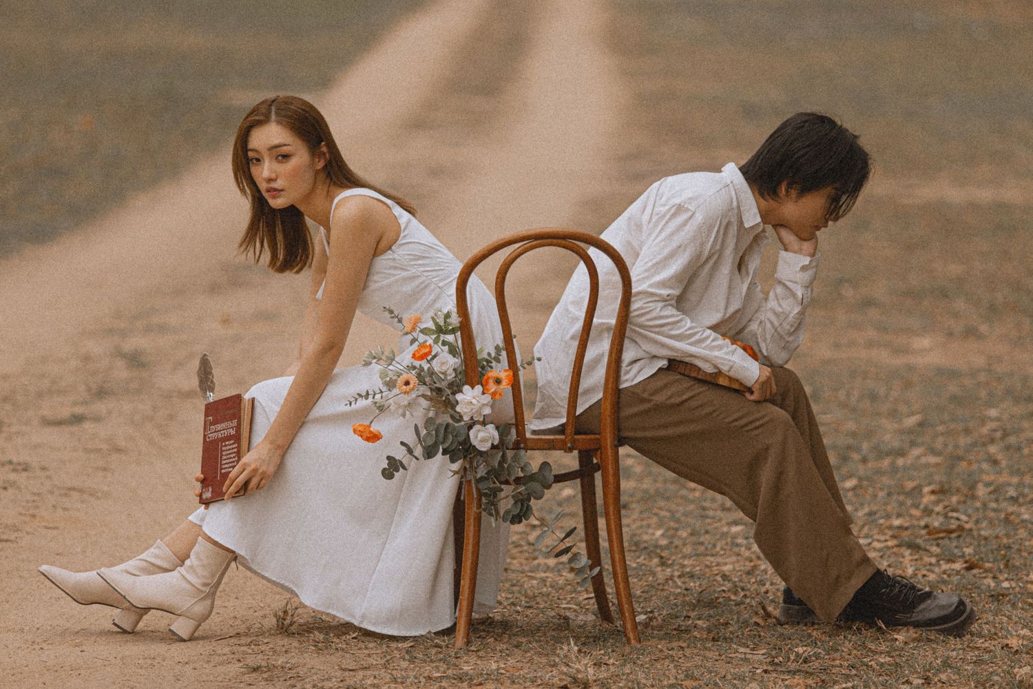 Asian couple in casual white and tan outfits sit back-to-back on a chair with flowers on a rustic dirt road.