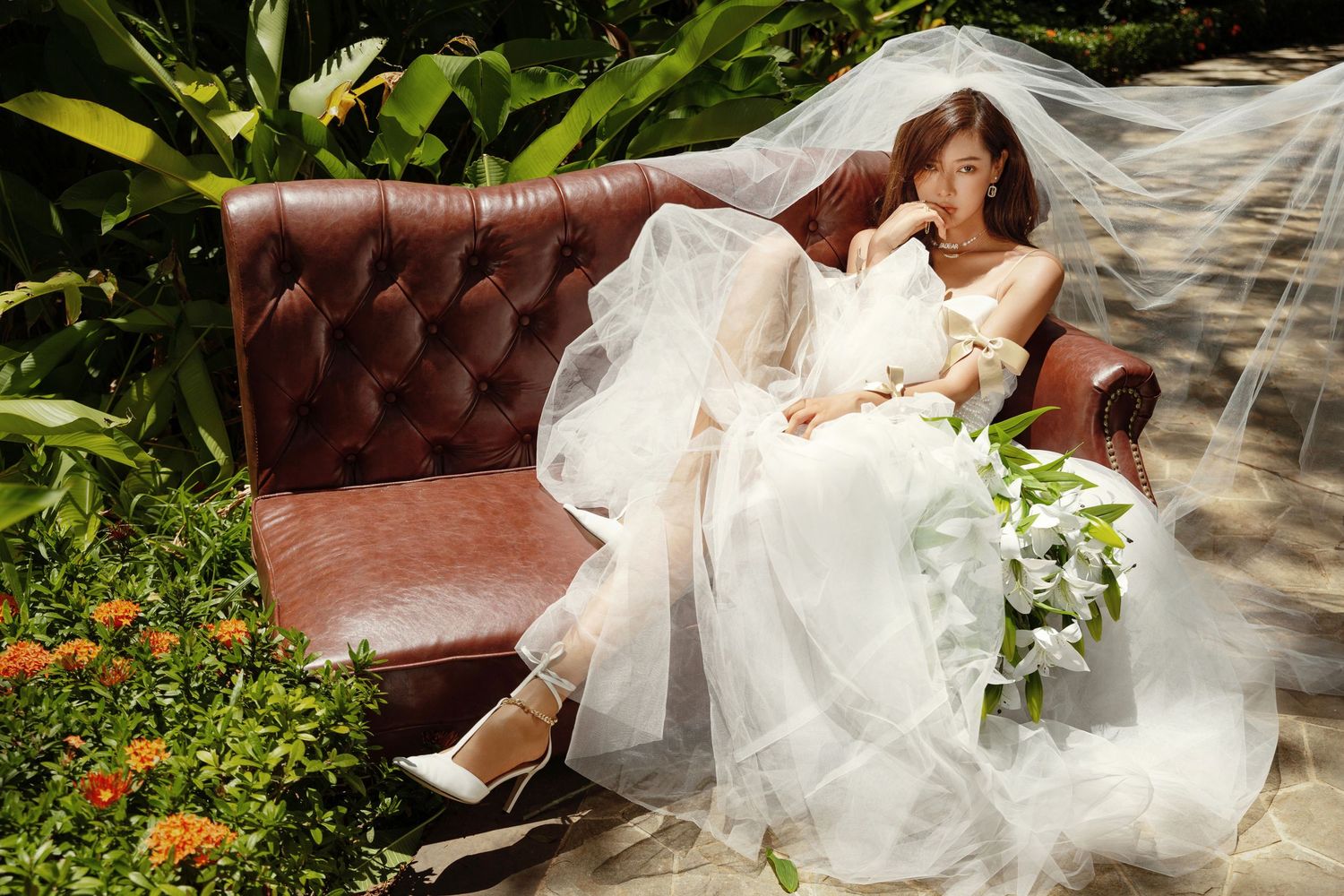 Beautiful bride in white wedding gown and veil relaxes on vintage leather sofa holding white flower bouquet outdoors.