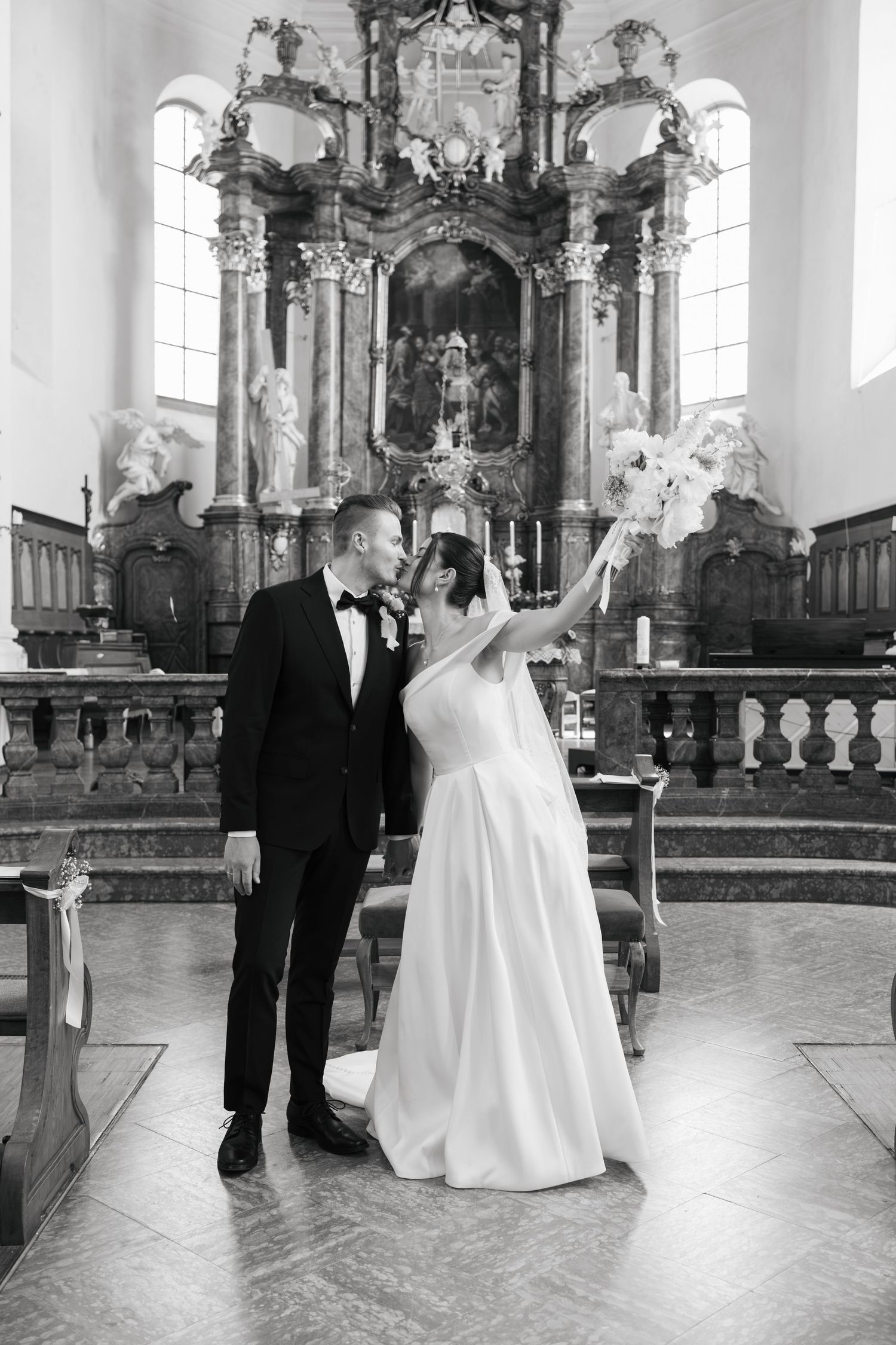 Black and white photo of bride and groom standing at church altar, bride raising bouquet, ornate baroque decor behind them.