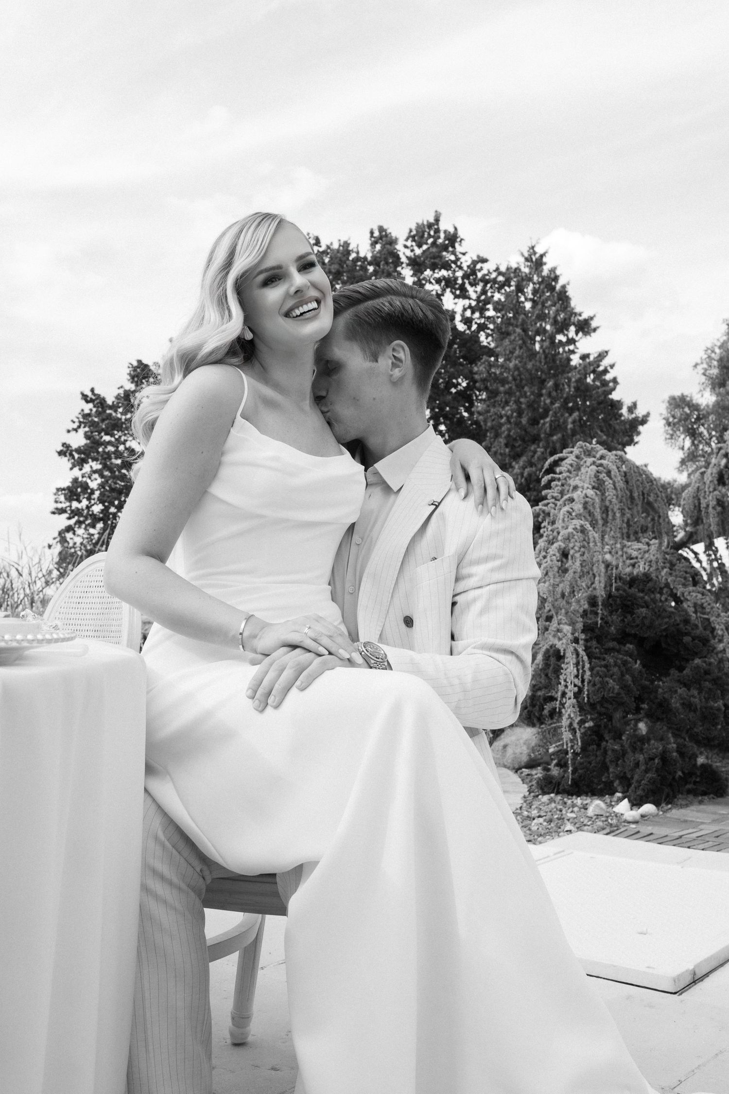 Black and white photo of a smiling bride in white dress with groom in suit sharing a tender moment outdoors.