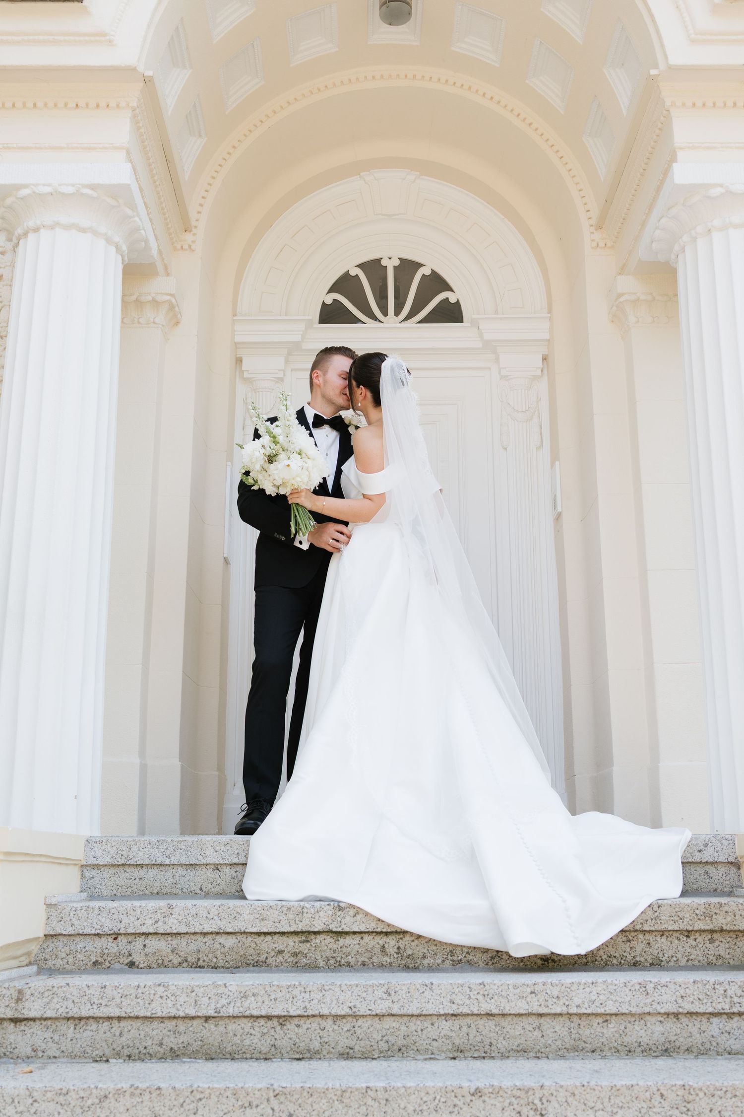 Bride and groom share a romantic kiss on marble steps before elegant white columns with arched doorway.