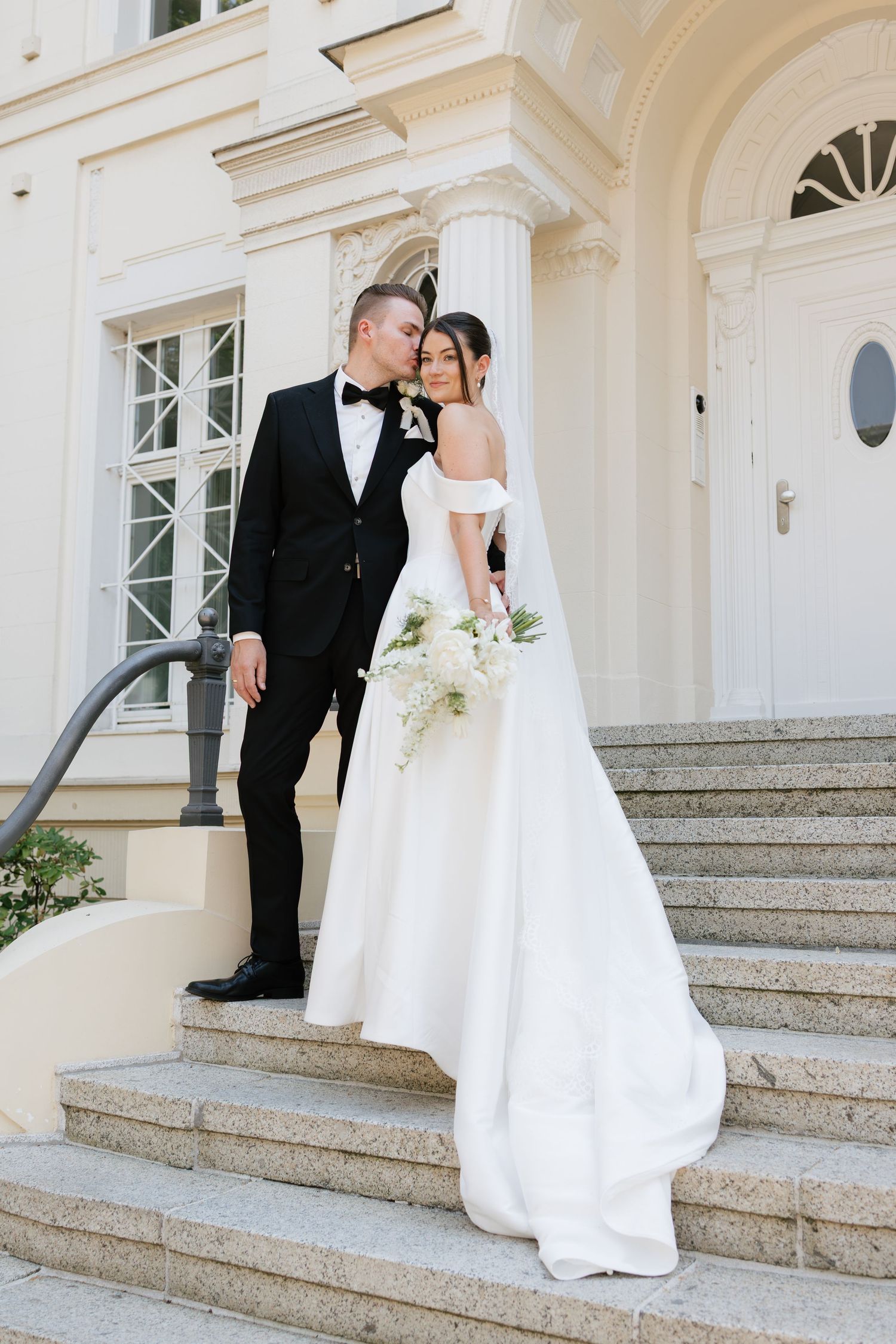 Elegant bride in off-shoulder white gown and groom in black tuxedo posing on stone steps of a classic white building.