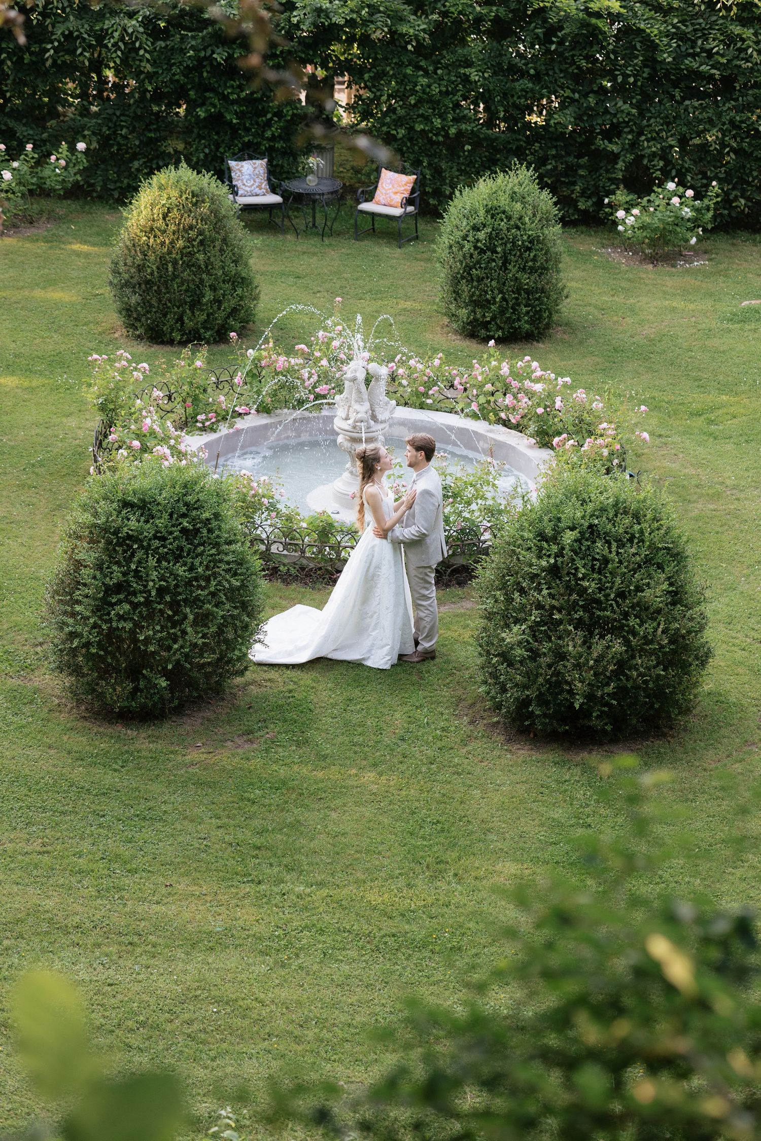 Bride and groom standing by a decorative fountain surrounded by trimmed hedges and roses in a lush garden.