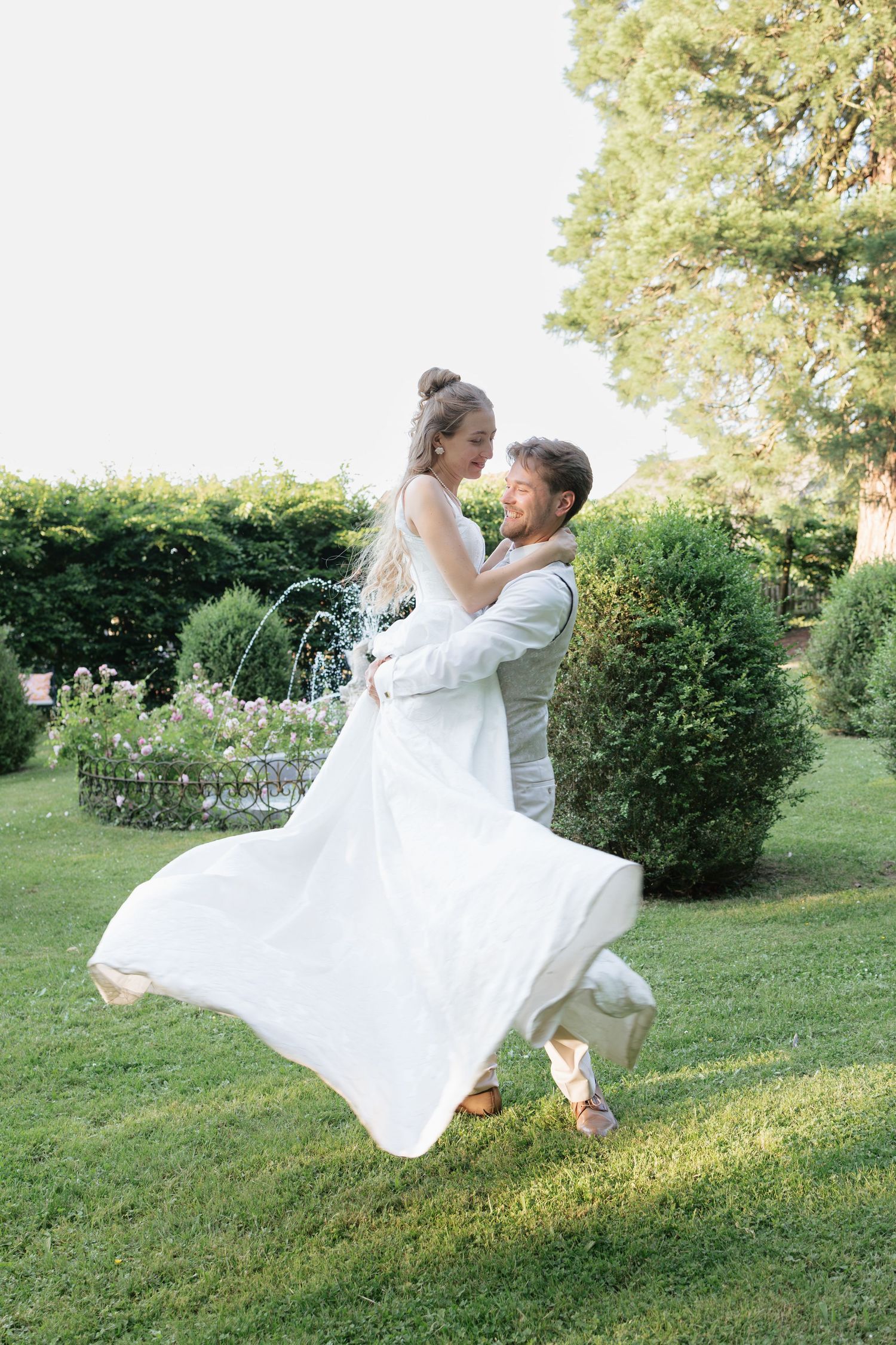 Groom lifting bride in white dress outdoors in lush garden, romantic wedding couple moment.