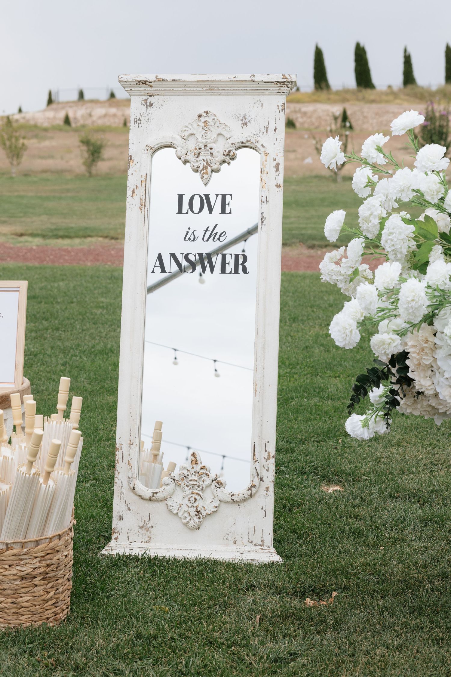 Vintage white ornate mirror with Love is the Answer text at outdoor wedding ceremony with white floral arrangements.