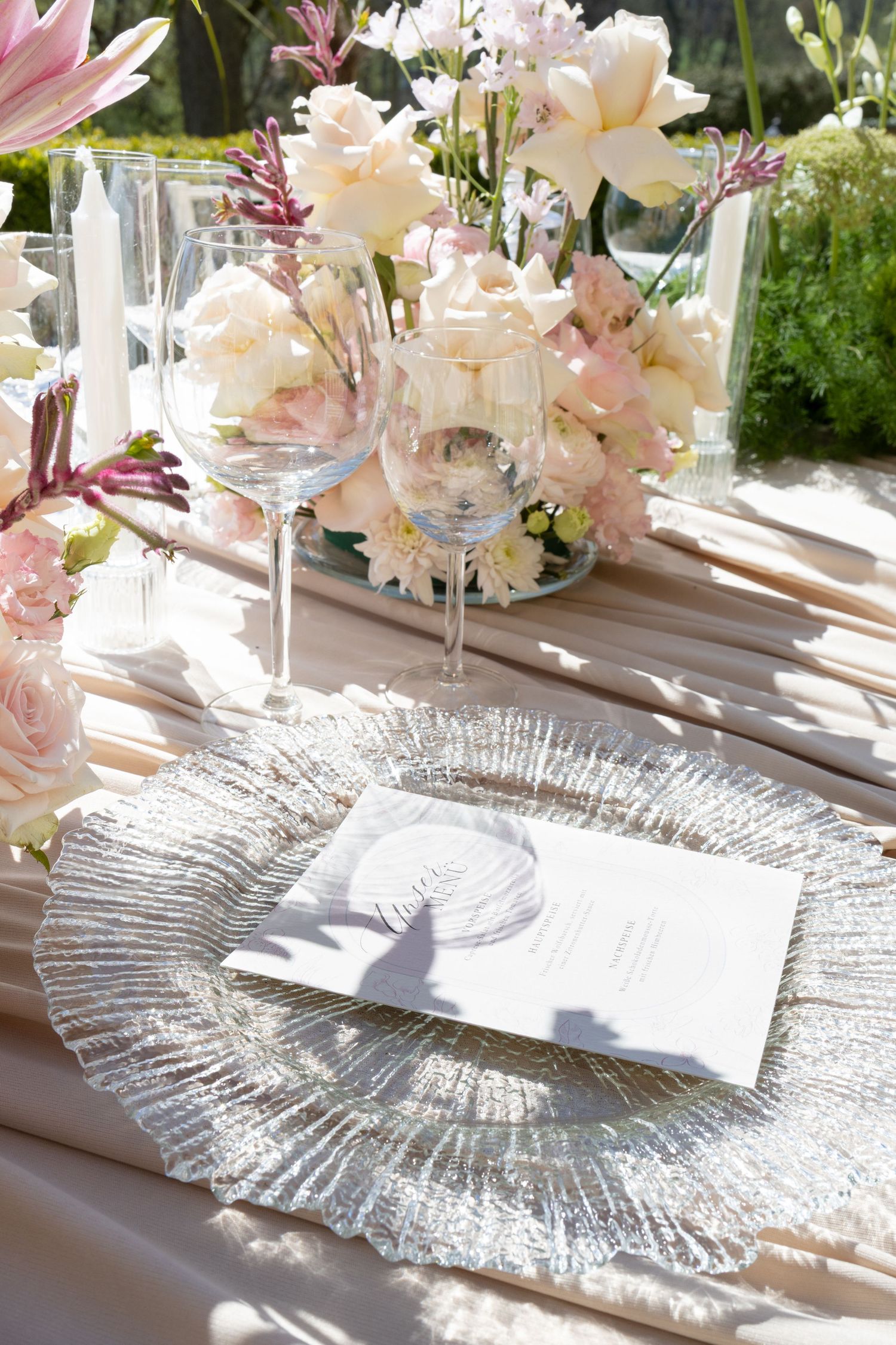 Elegant wedding table setting with crystal charger plate, wine glasses, and blush floral centerpiece in soft natural light.