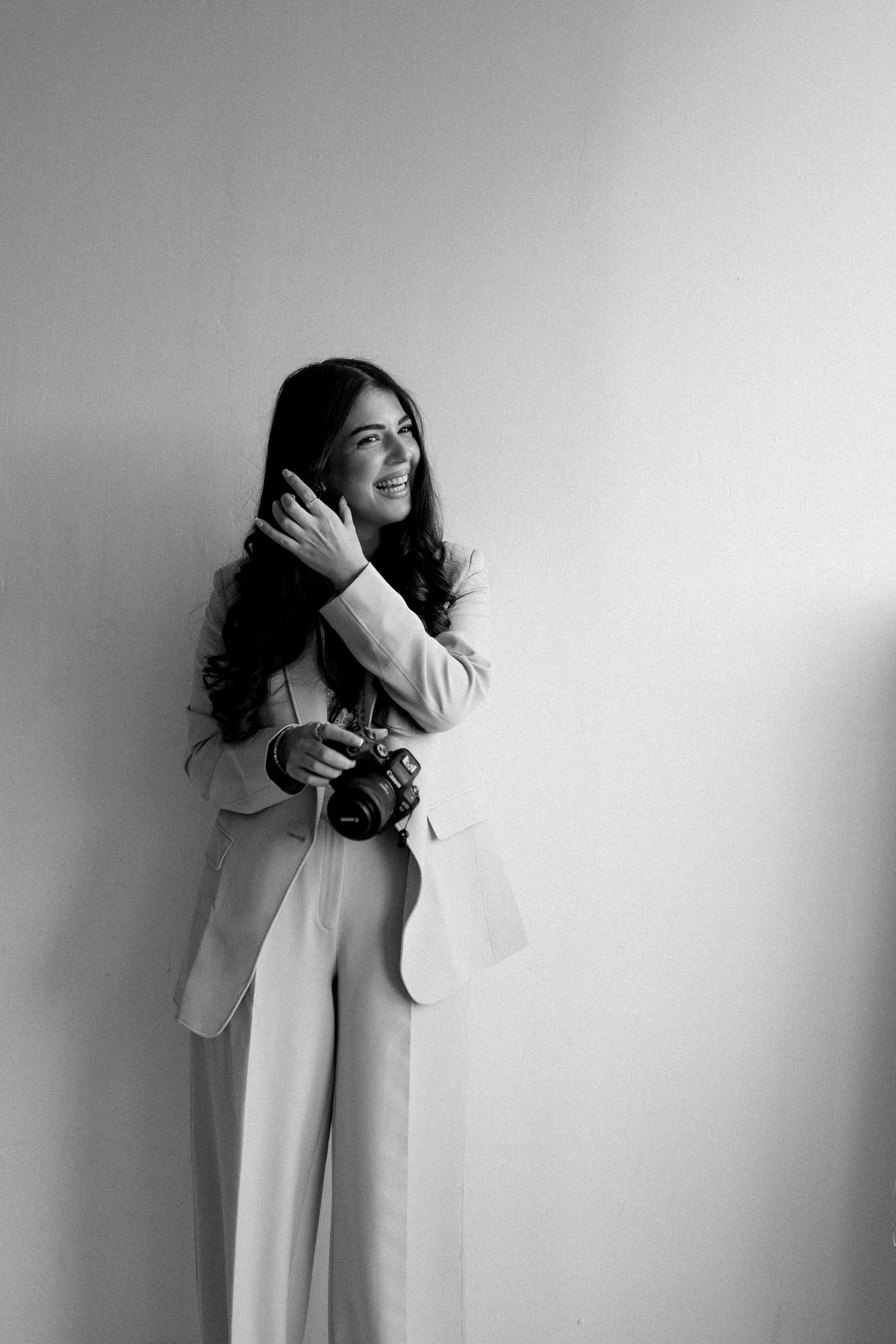 Smiling woman in light blazer holding a camera while leaning against a white wall in black and white photo.