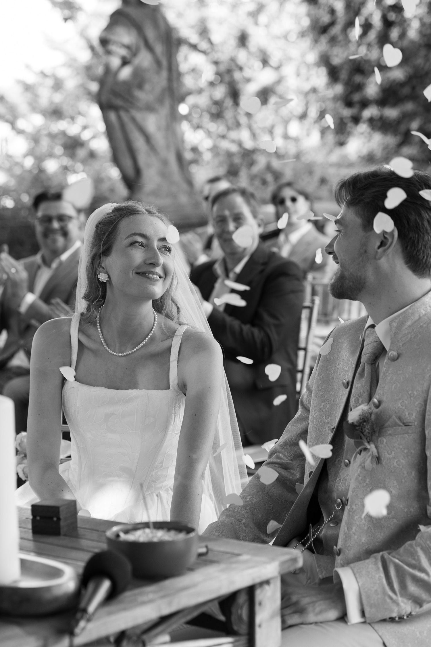 Black and white photo of smiling bride and groom at outdoor wedding ceremony with guests and flower petals in background.
