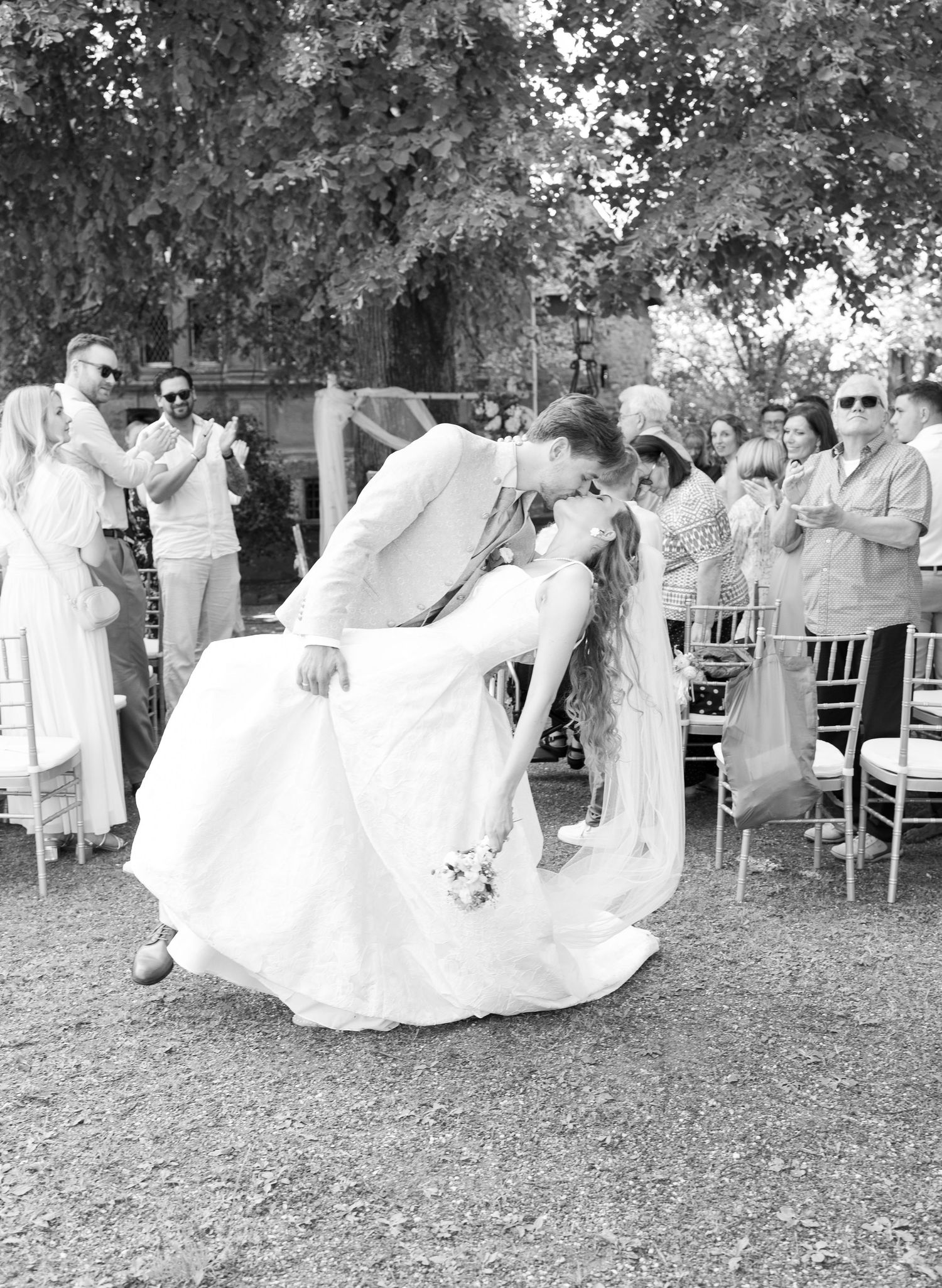 Black and white photo of groom dipping bride for a kiss at outdoor wedding ceremony with guests applauding.