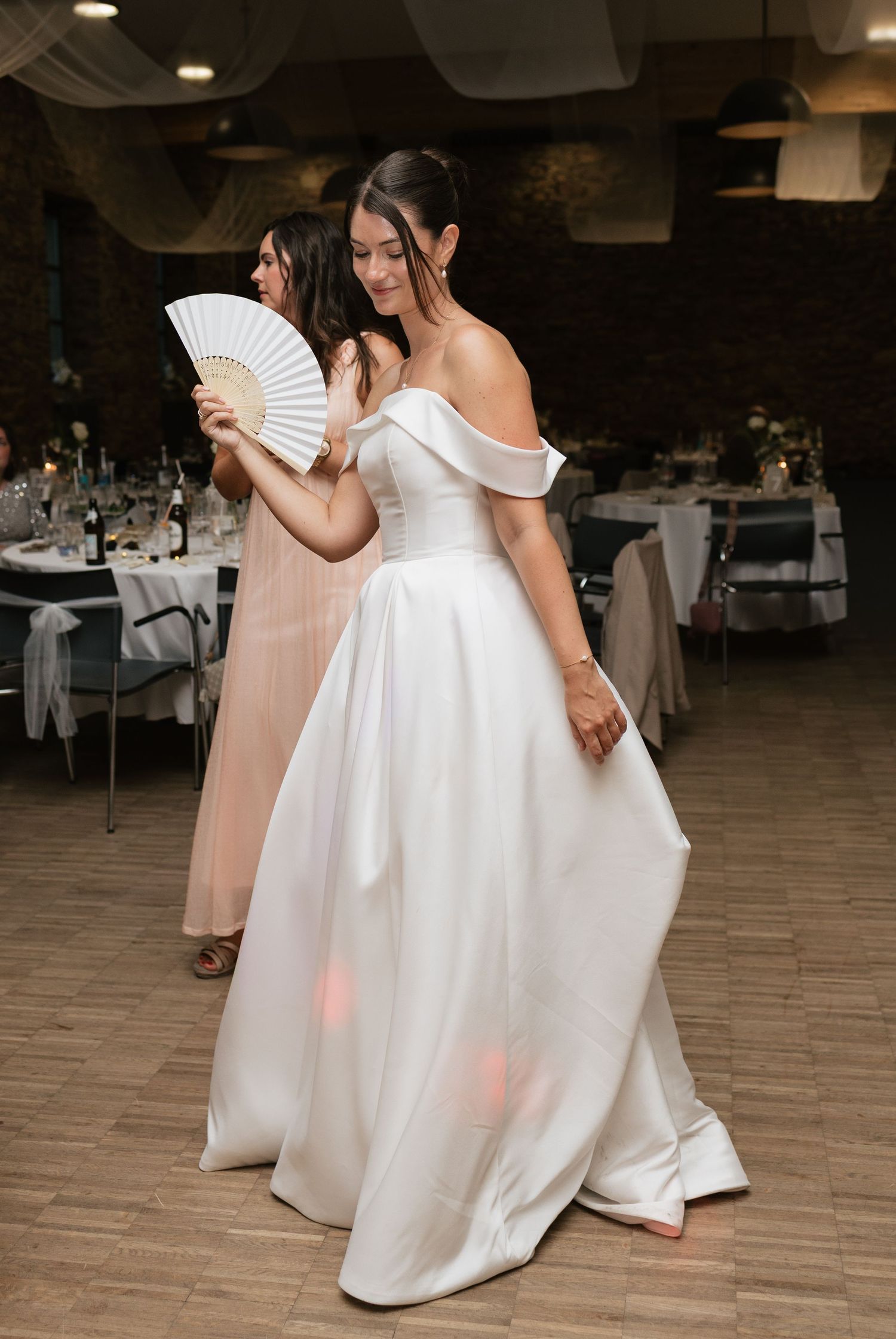 Bride in off-shoulder white ball gown dancing with a hand fan at elegant wedding reception.