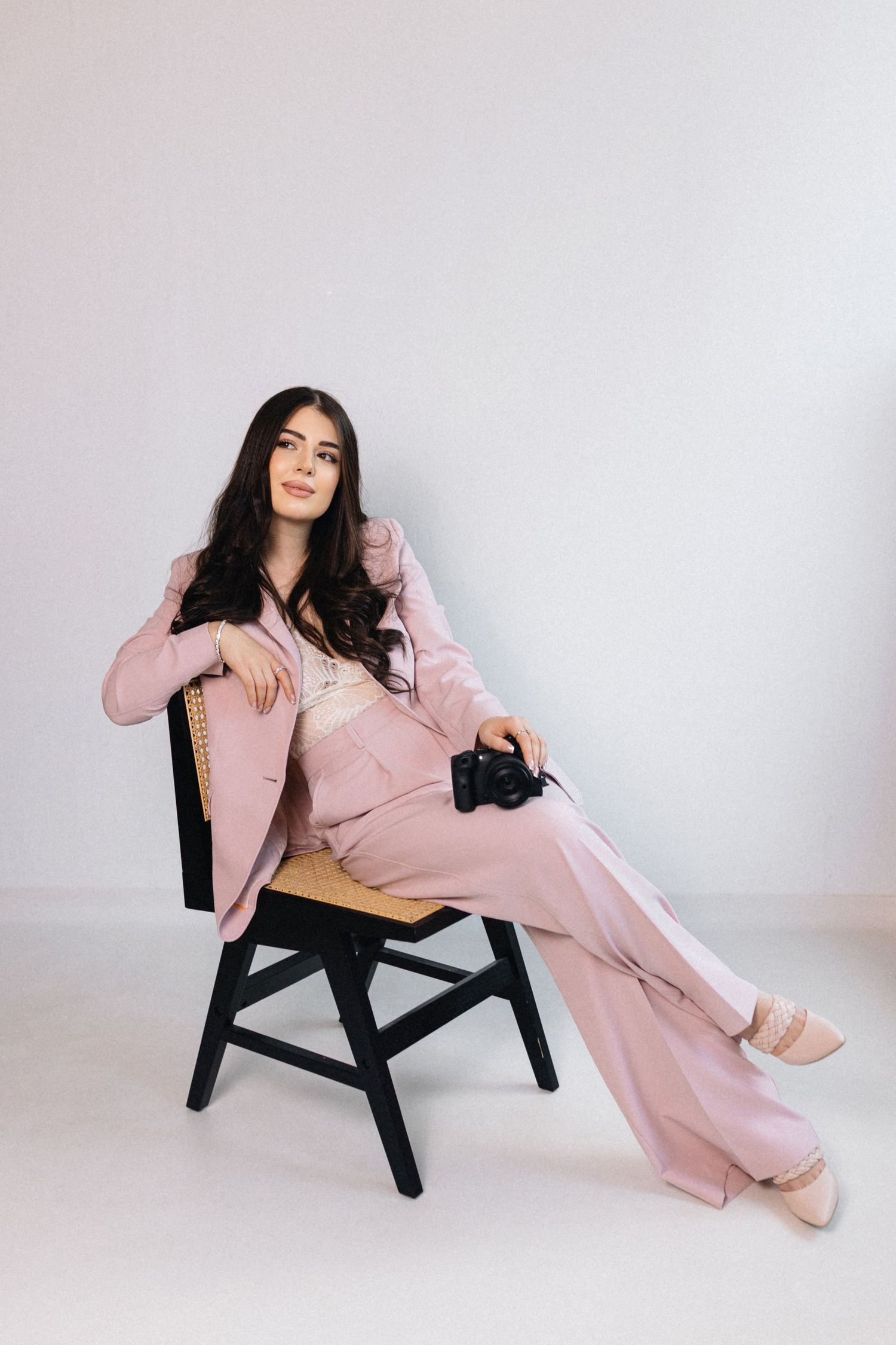 Woman in pink suit sitting on black chair holding camera against white background.