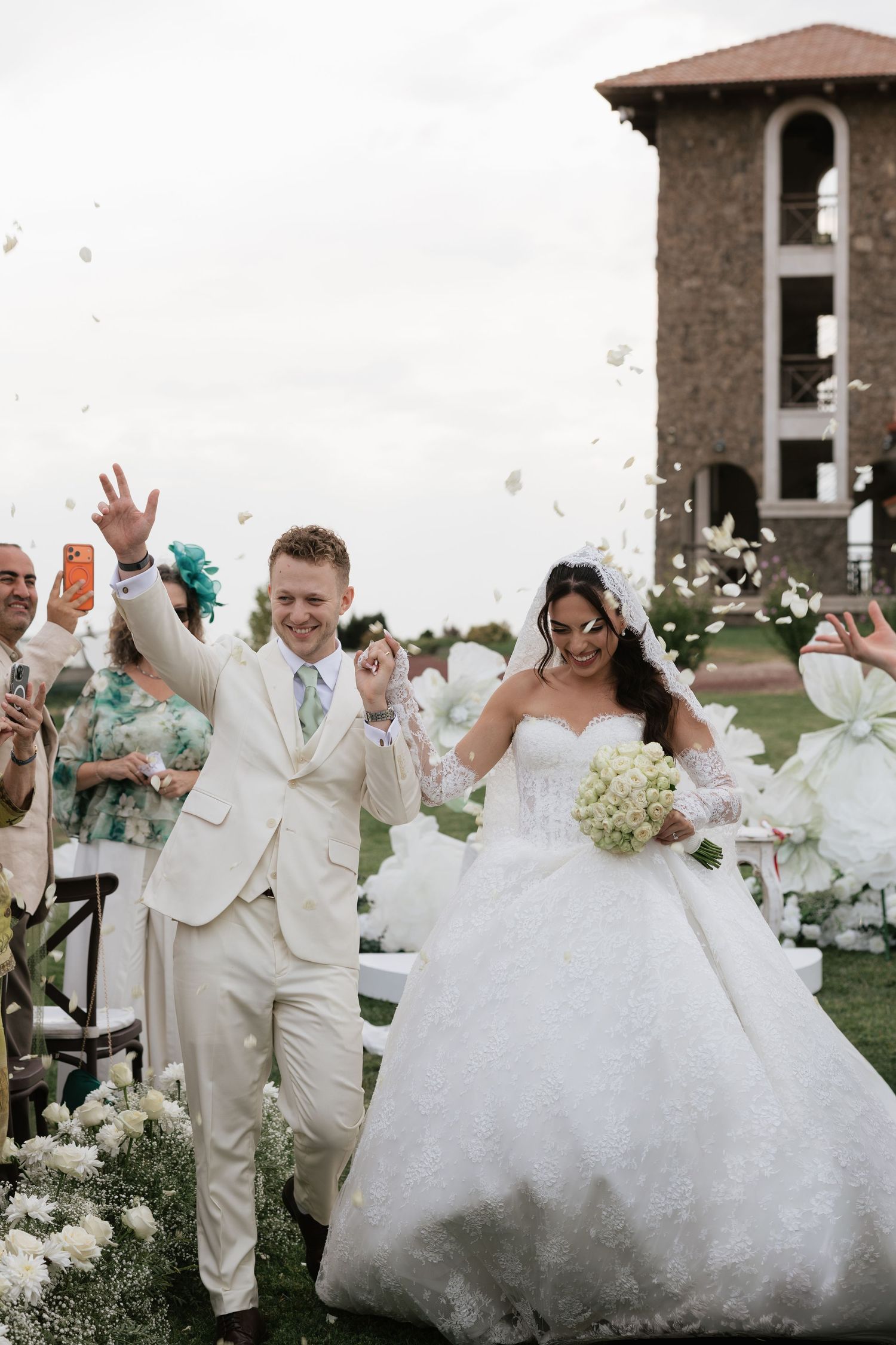 Bride and groom walk down the aisle as guests toss white flower petals at an outdoor wedding ceremony.