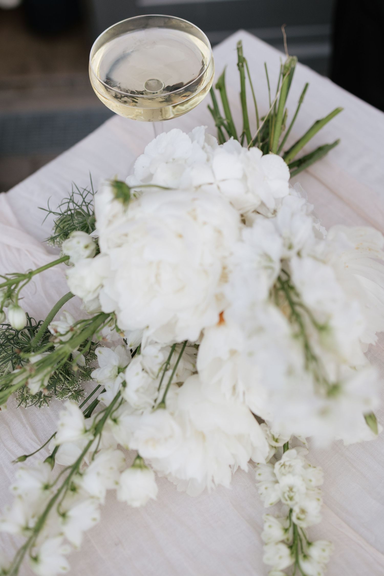 White peony bridal bouquet with greenery beside a glass of champagne on a white table.