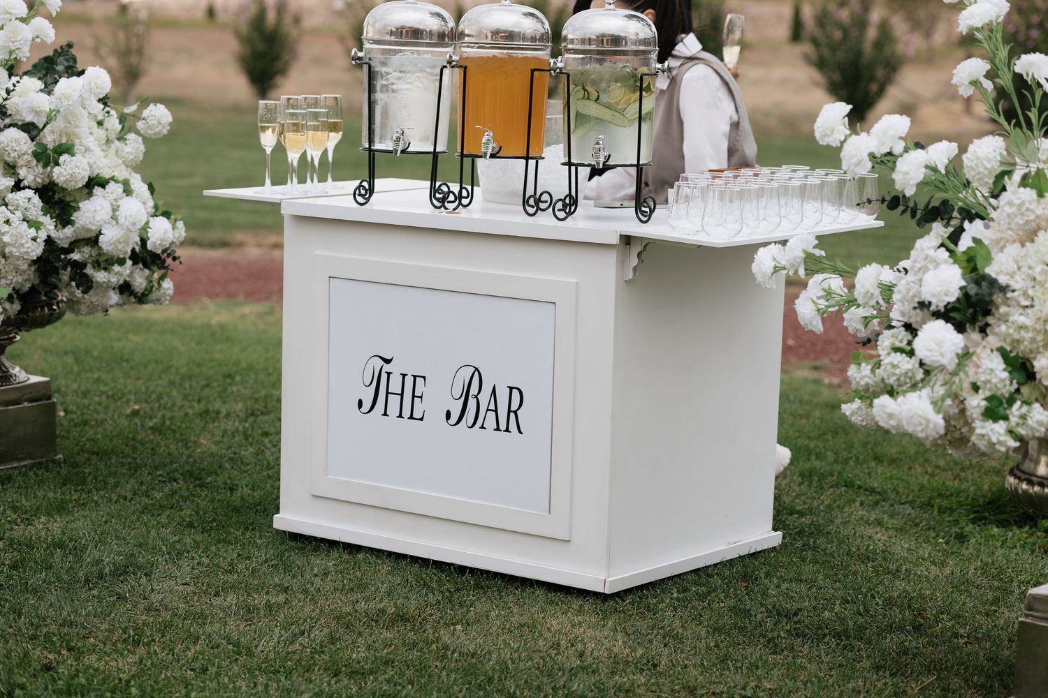 Elegant white outdoor wedding bar cart with beverage dispensers, champagne flutes, and white floral arrangements on green lawn.