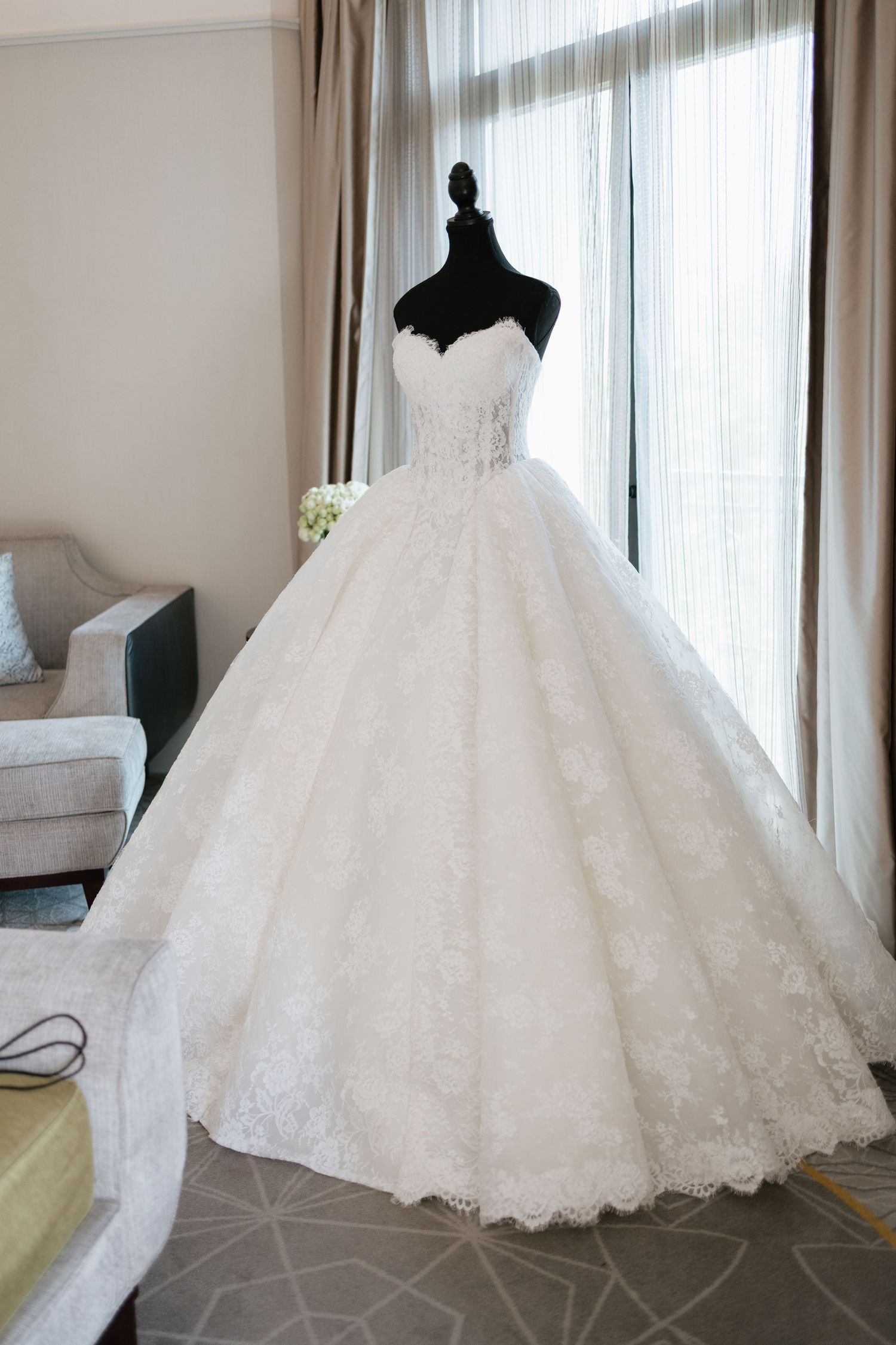 Elegant strapless white lace ball gown wedding dress displayed on a black mannequin near a bright window.