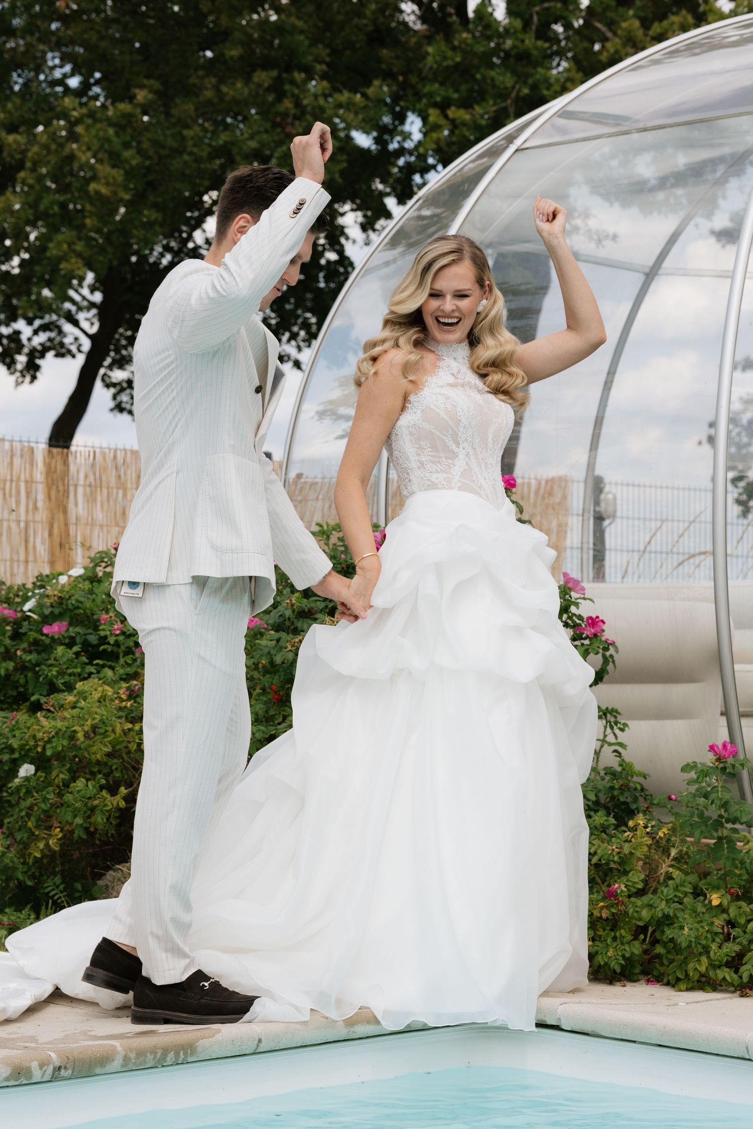 Joyful bride and groom celebrating in white wedding attire by a pool, raising arms in excitement outdoors.