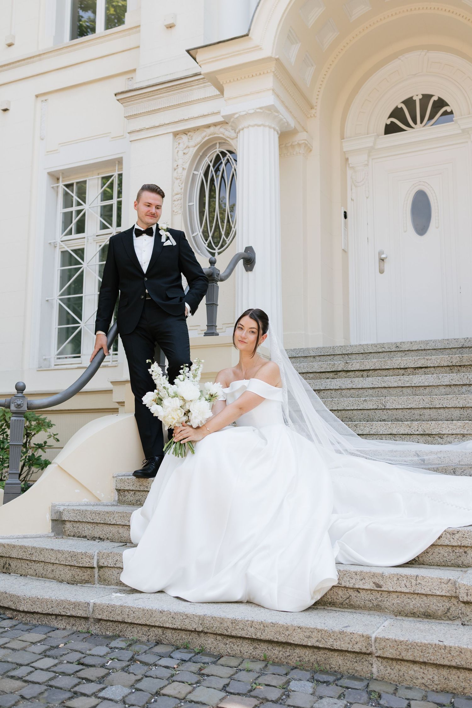 Bride in white gown sits on steps holding bouquet while groom in black tuxedo stands beside elegant building.