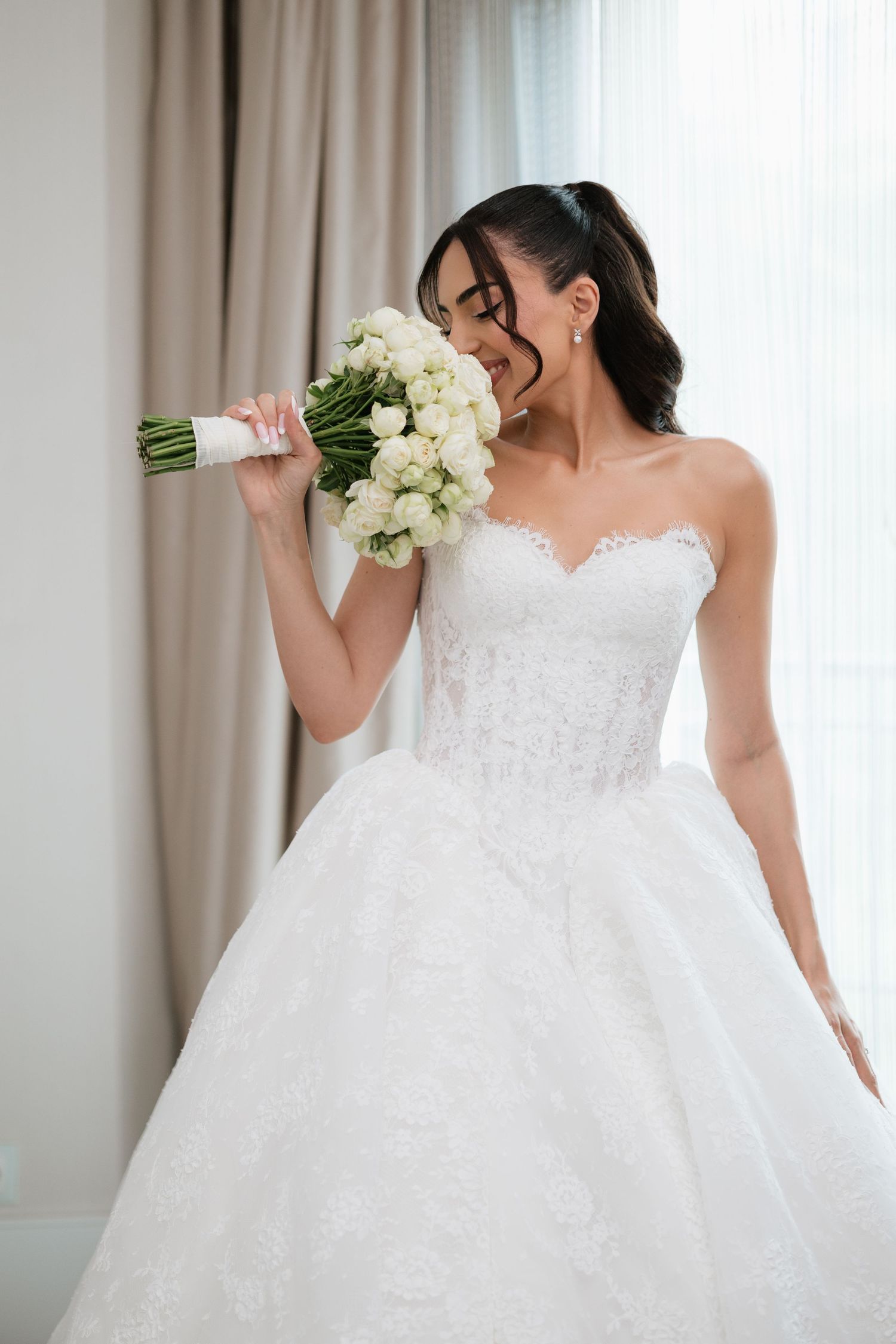 Bride in strapless lace ballgown smelling white rose bouquet by window on wedding day.