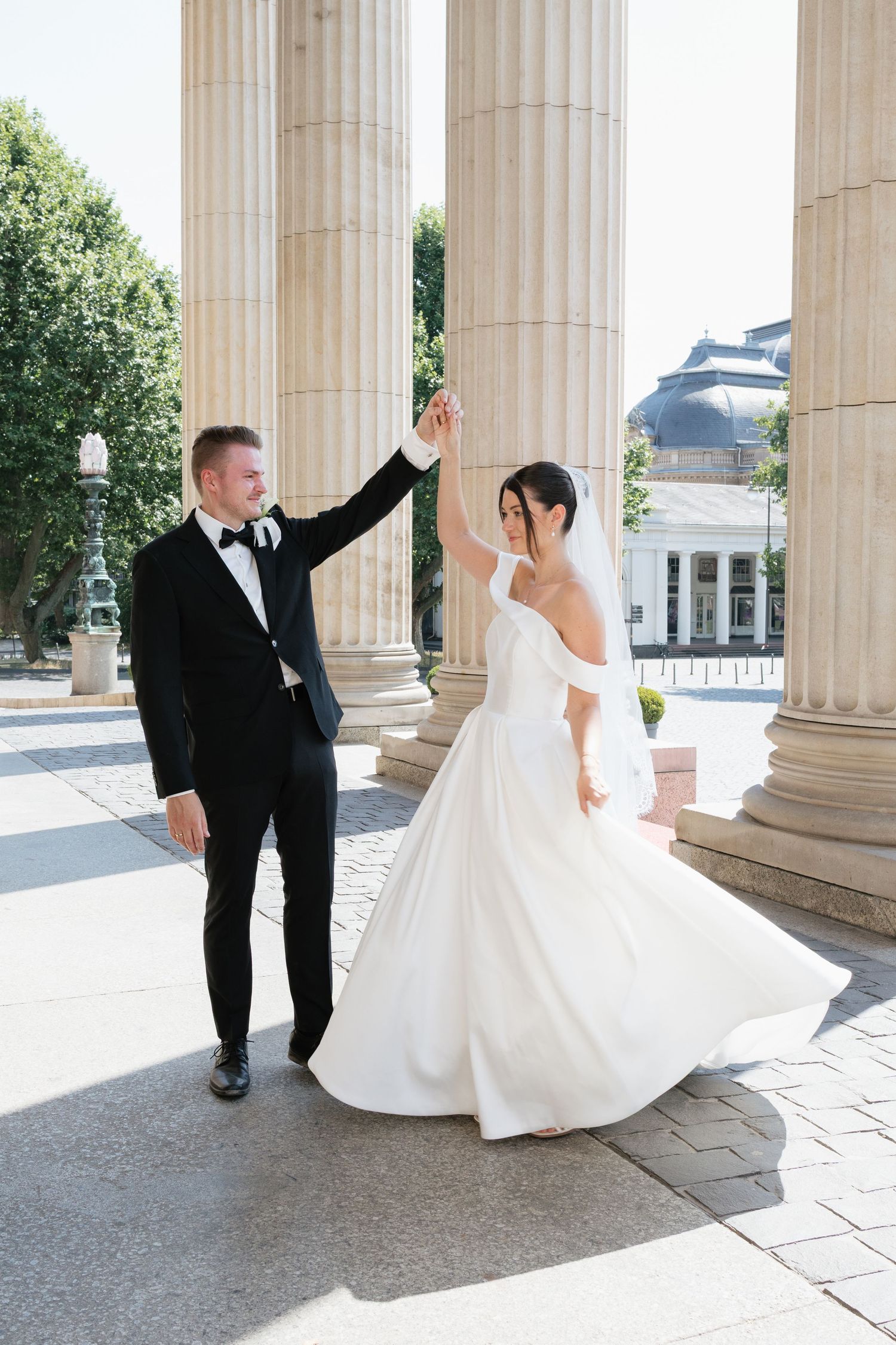 A groom in a black tuxedo twirls his bride in a white off-shoulder gown near grand columns outdoors.