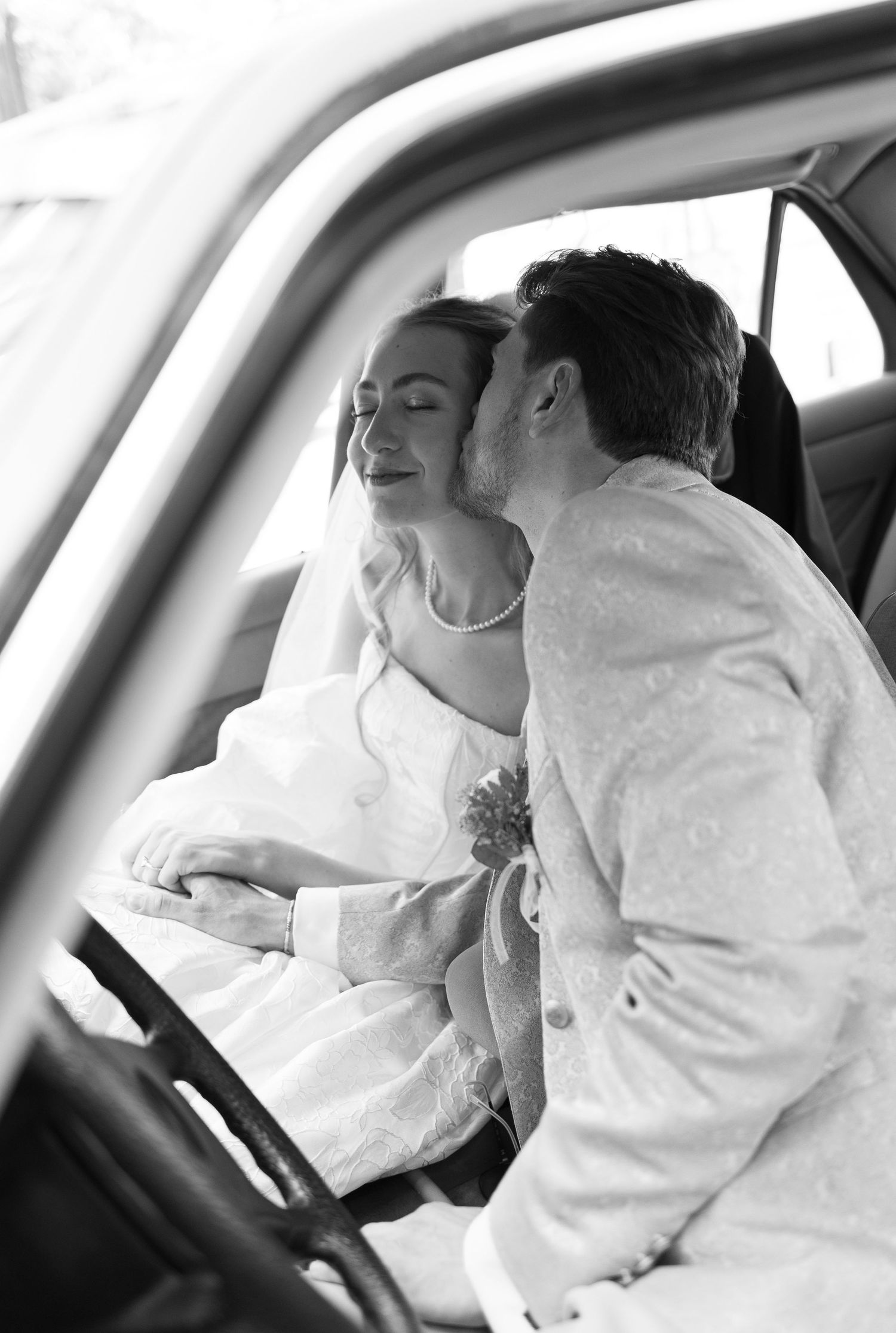 Black and white photo of a groom kissing bride on cheek in car on wedding day.