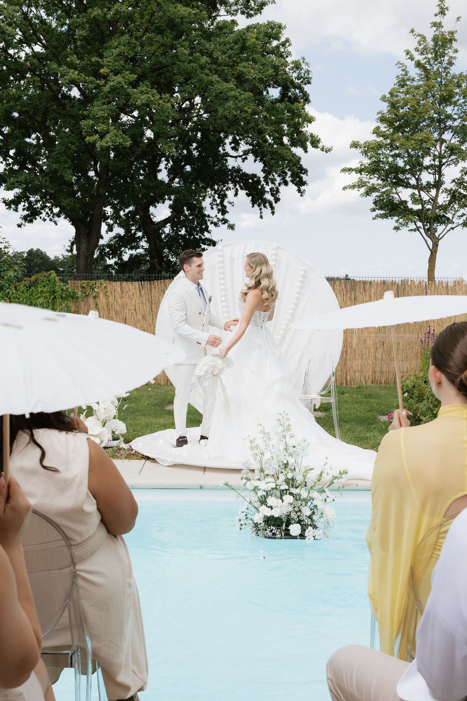 Outdoor wedding ceremony by a pool with bride and groom at white floral altar, guests seated under parasols.