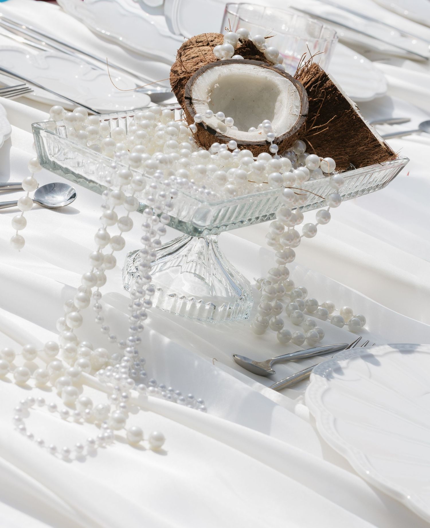 Elegant white table setting with pearl necklaces draped over a glass stand holding a halved coconut.