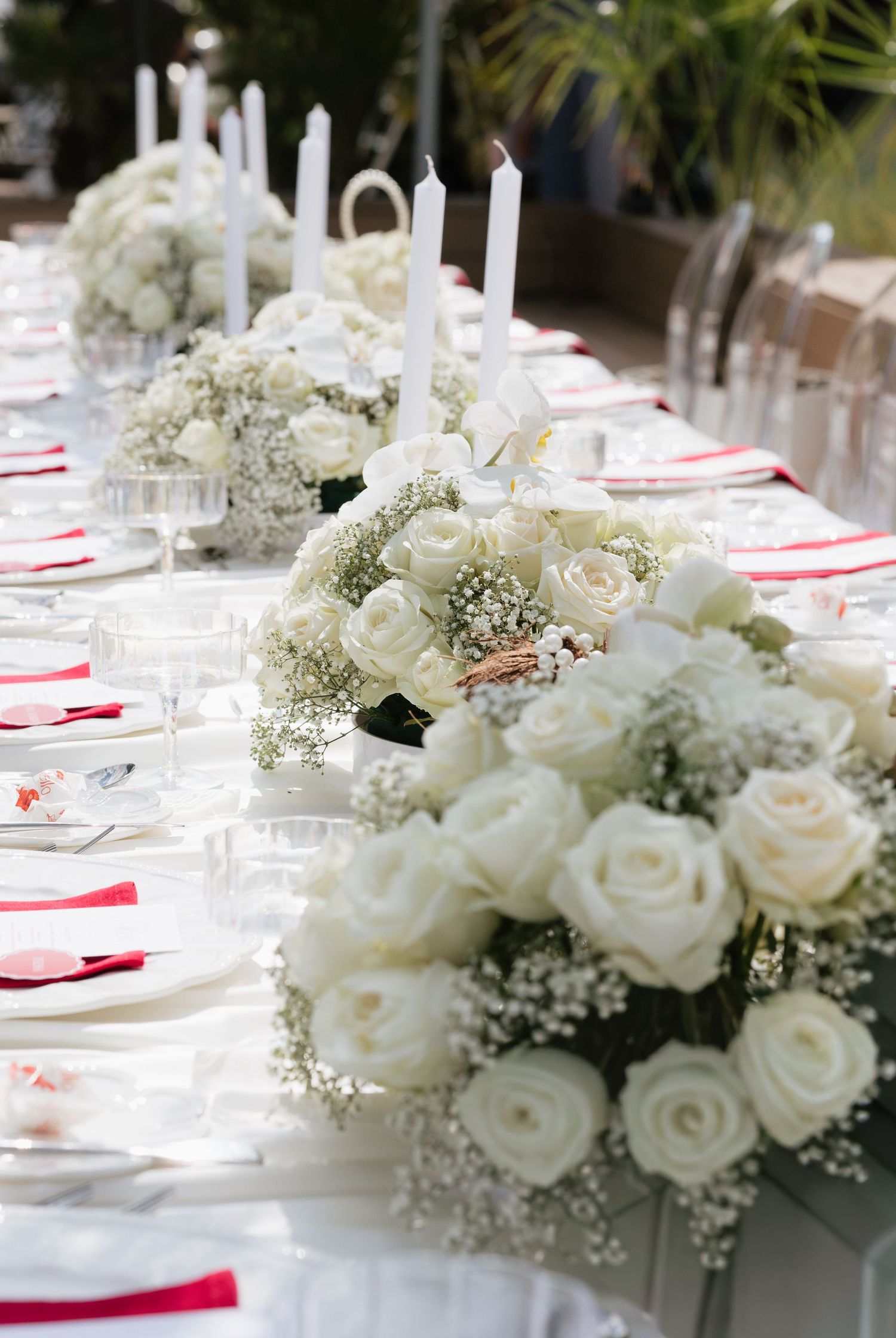 Elegant wedding table with white rose centerpieces, baby's breath, tall candles, and red napkins on a white tablecloth.